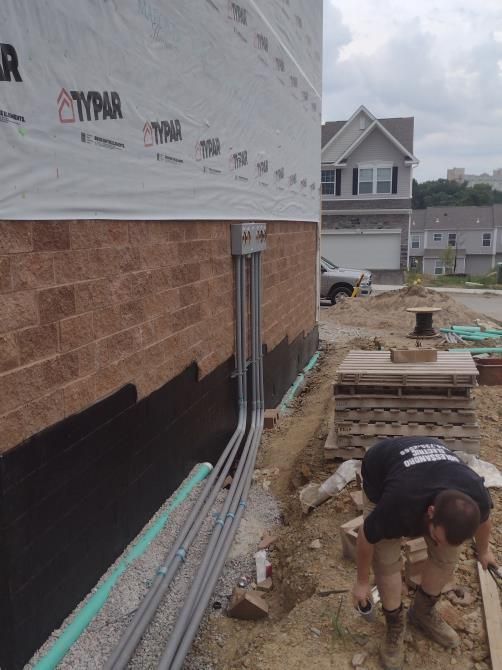 Construction worker digging beside a house foundation with exposed pipes and stacked materials.
