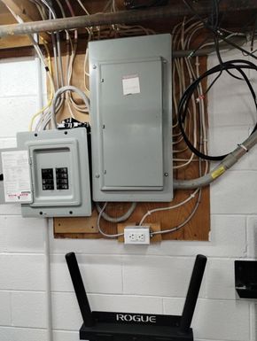 Electrical panels and exposed wiring mounted on a basement wall above a Rogue exercise rack.