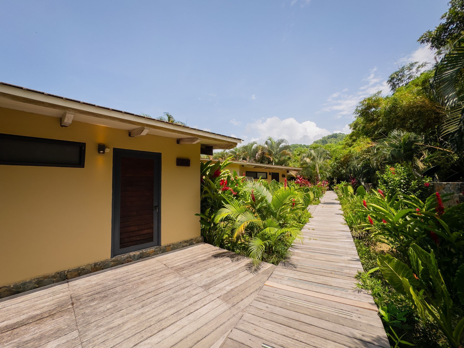 Wooden pathway between yellow buildings and lush greenery under a blue sky.