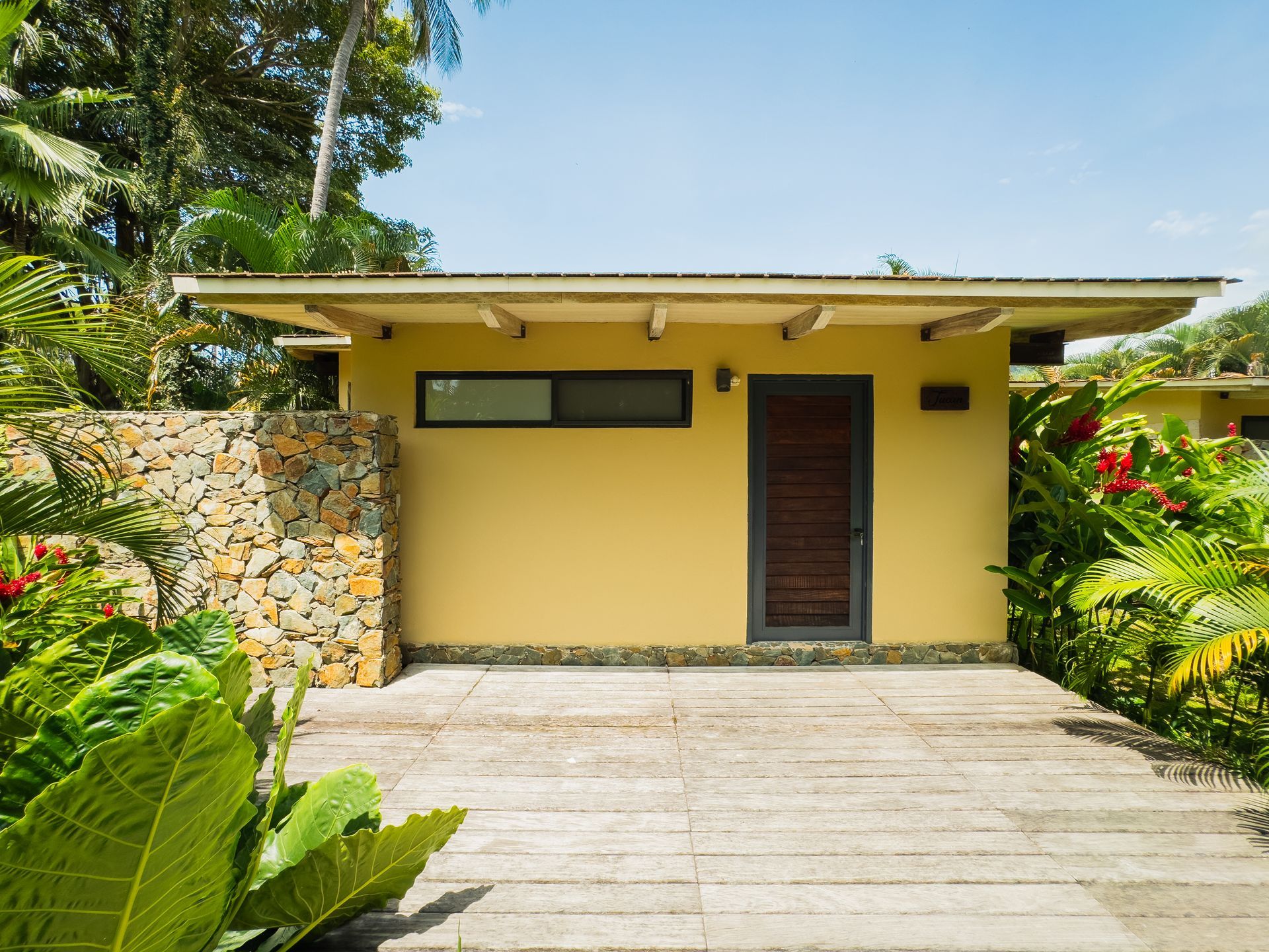 Yellow building with dark door and stone wall, surrounded by lush green plants and wooden pathway.