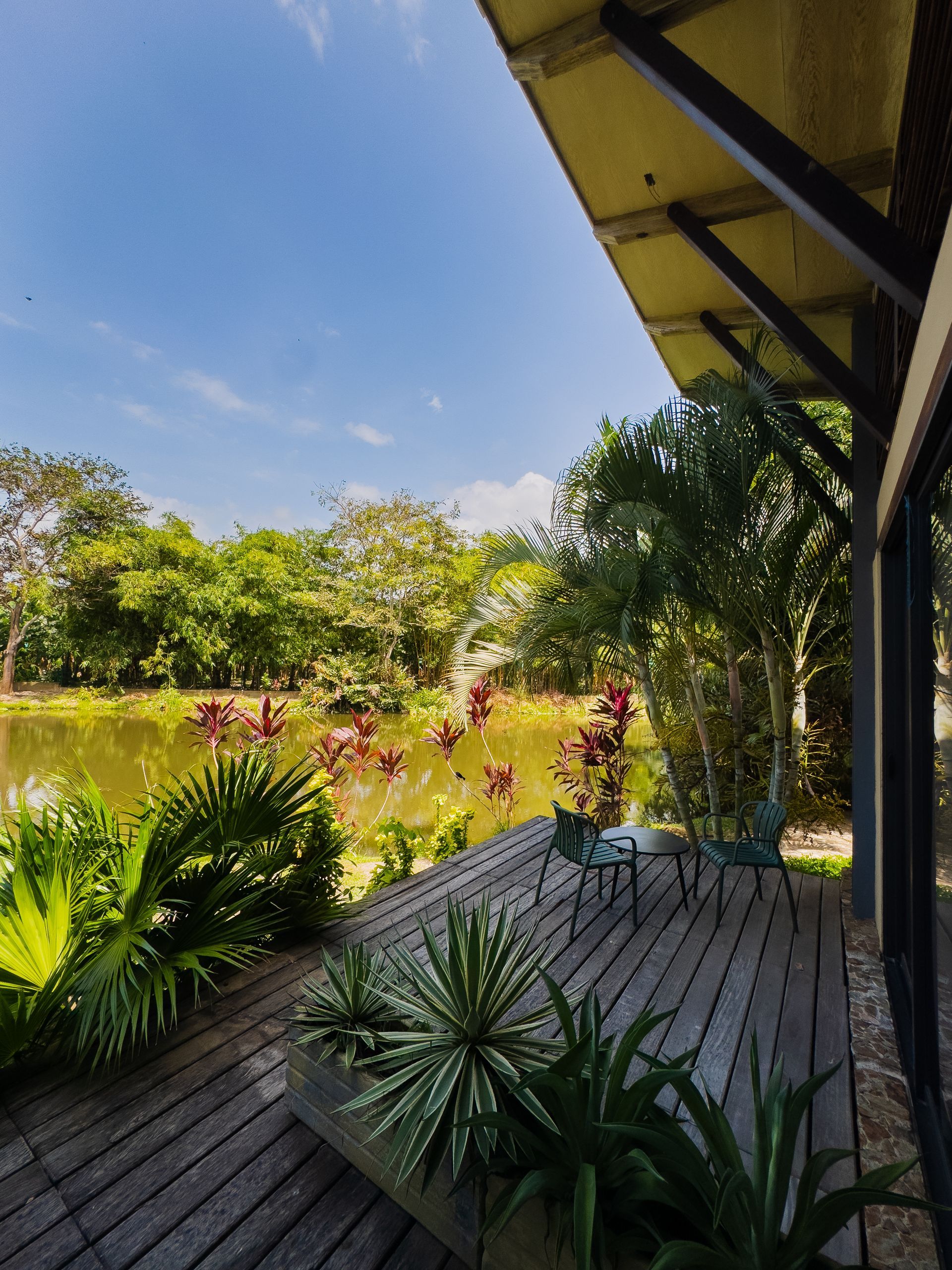 Wooden deck overlooking a pond, with lush greenery and a small table and chairs under a clear blue sky.