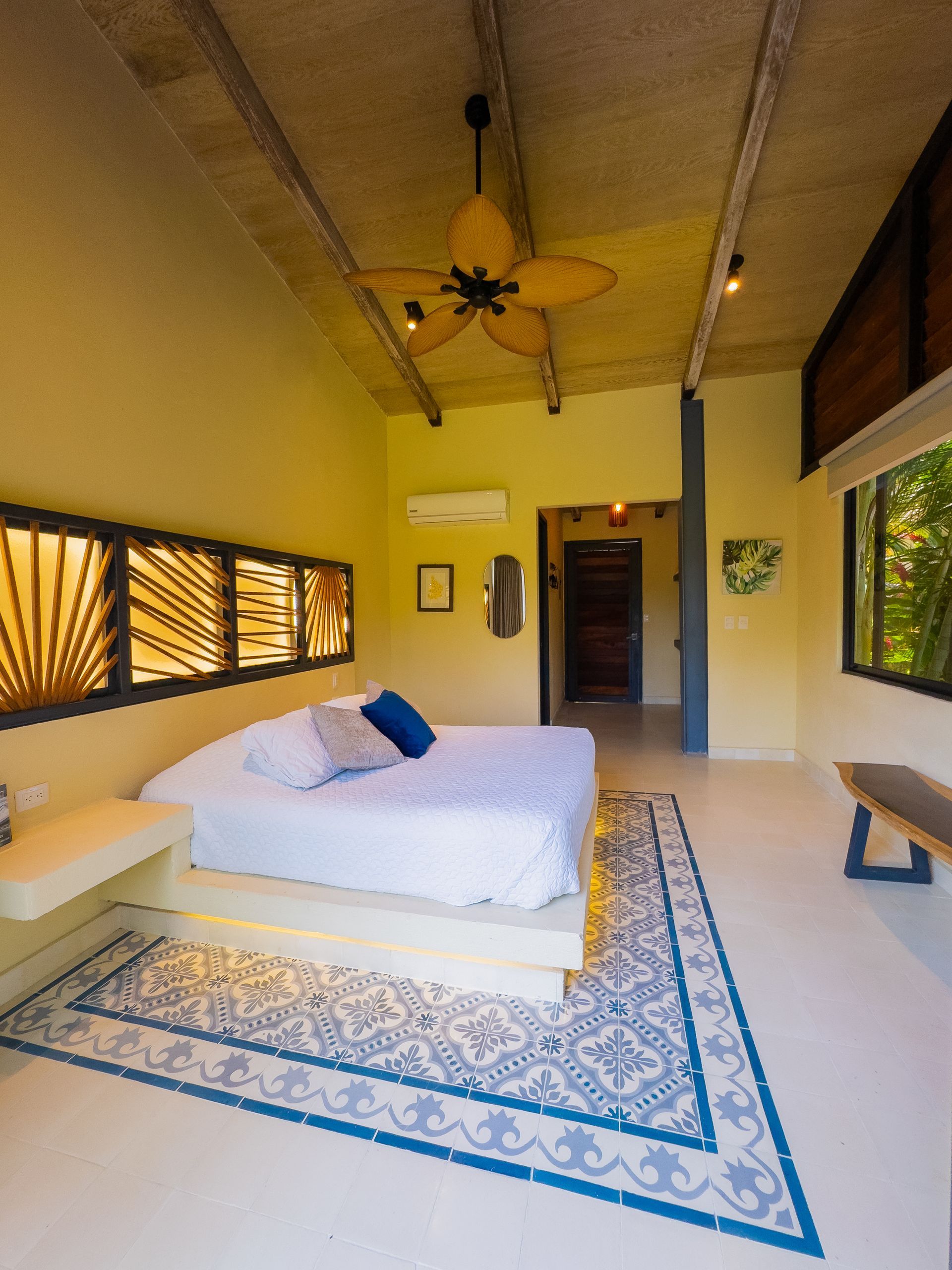 Bedroom with white bed, patterned rug, wooden beams, and a fan.