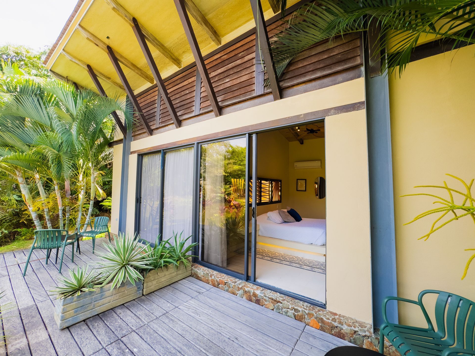 Wooden deck with potted plants, leads to a room with sliding glass doors. Yellow roof overhang. Green chairs.