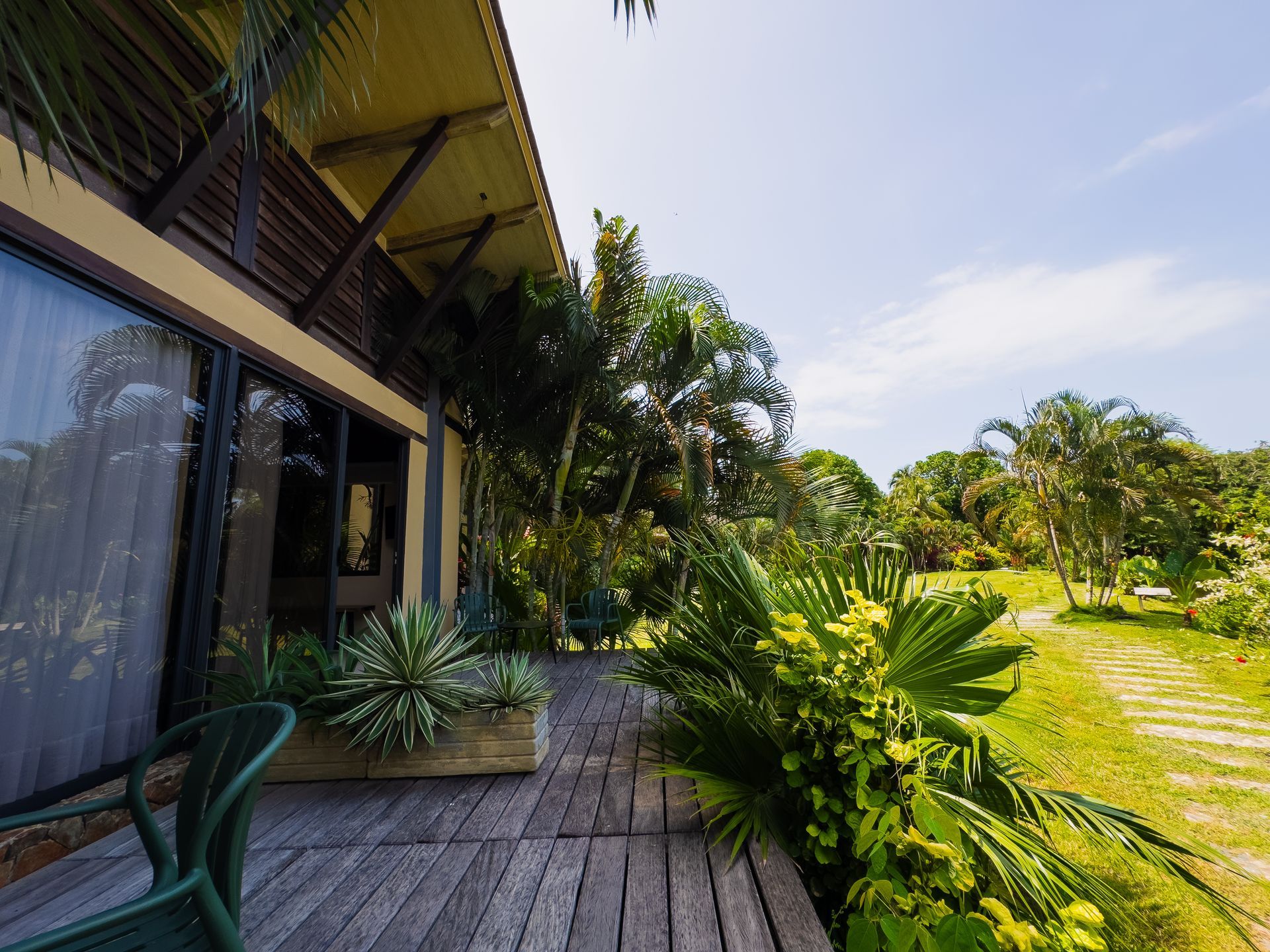 Wooden deck with large windows, lush greenery, and a path leading into the distance under a bright sky.