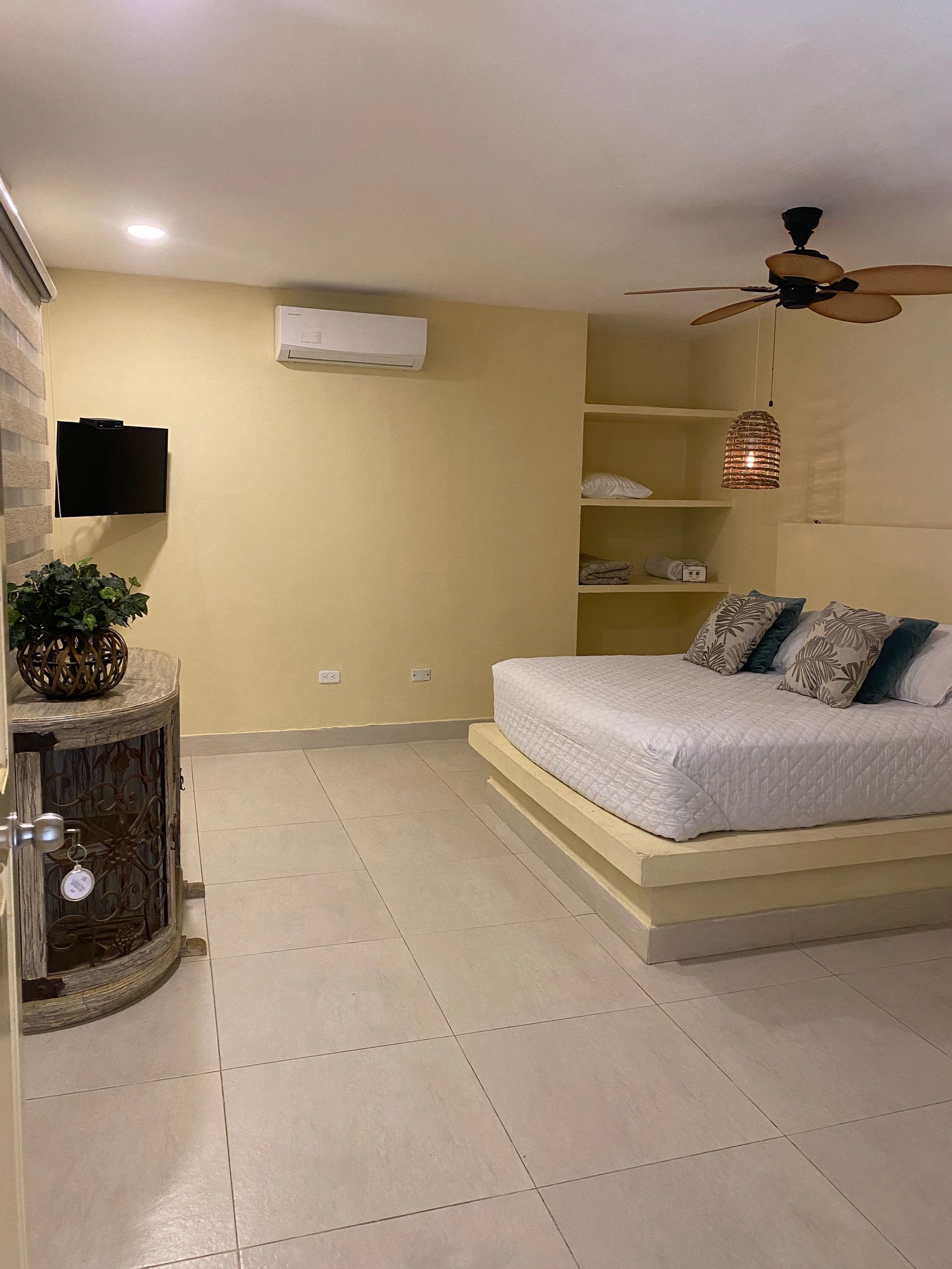 Bedroom with bed, shelves, TV, and air conditioner. Beige walls, tile floor.
