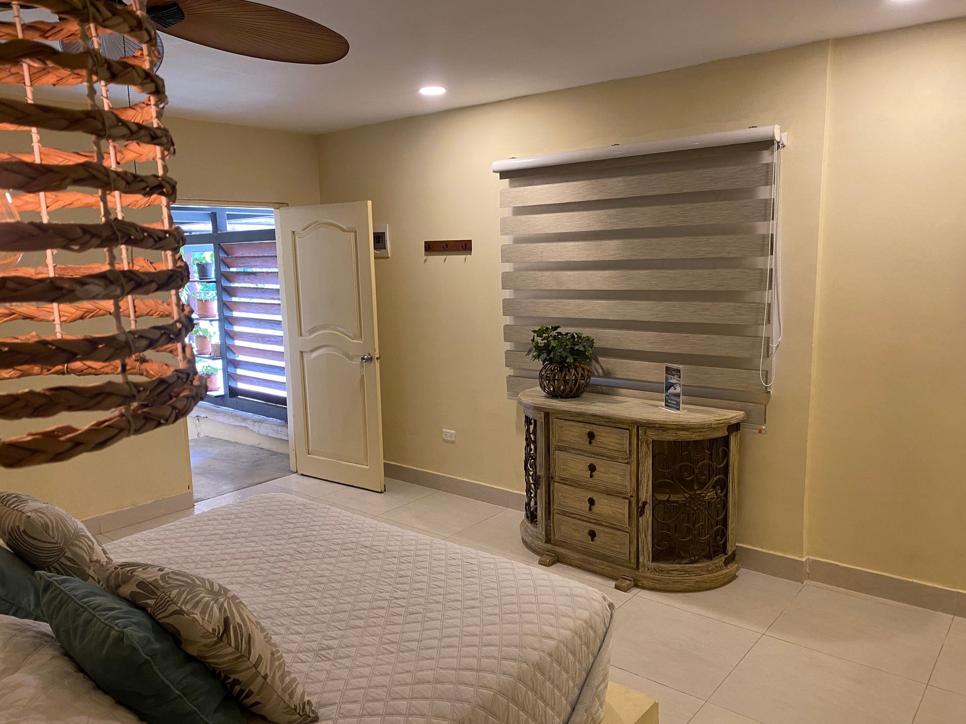 Bedroom with a bed, dresser, window with blinds, and open door to a patio. Light beige walls.