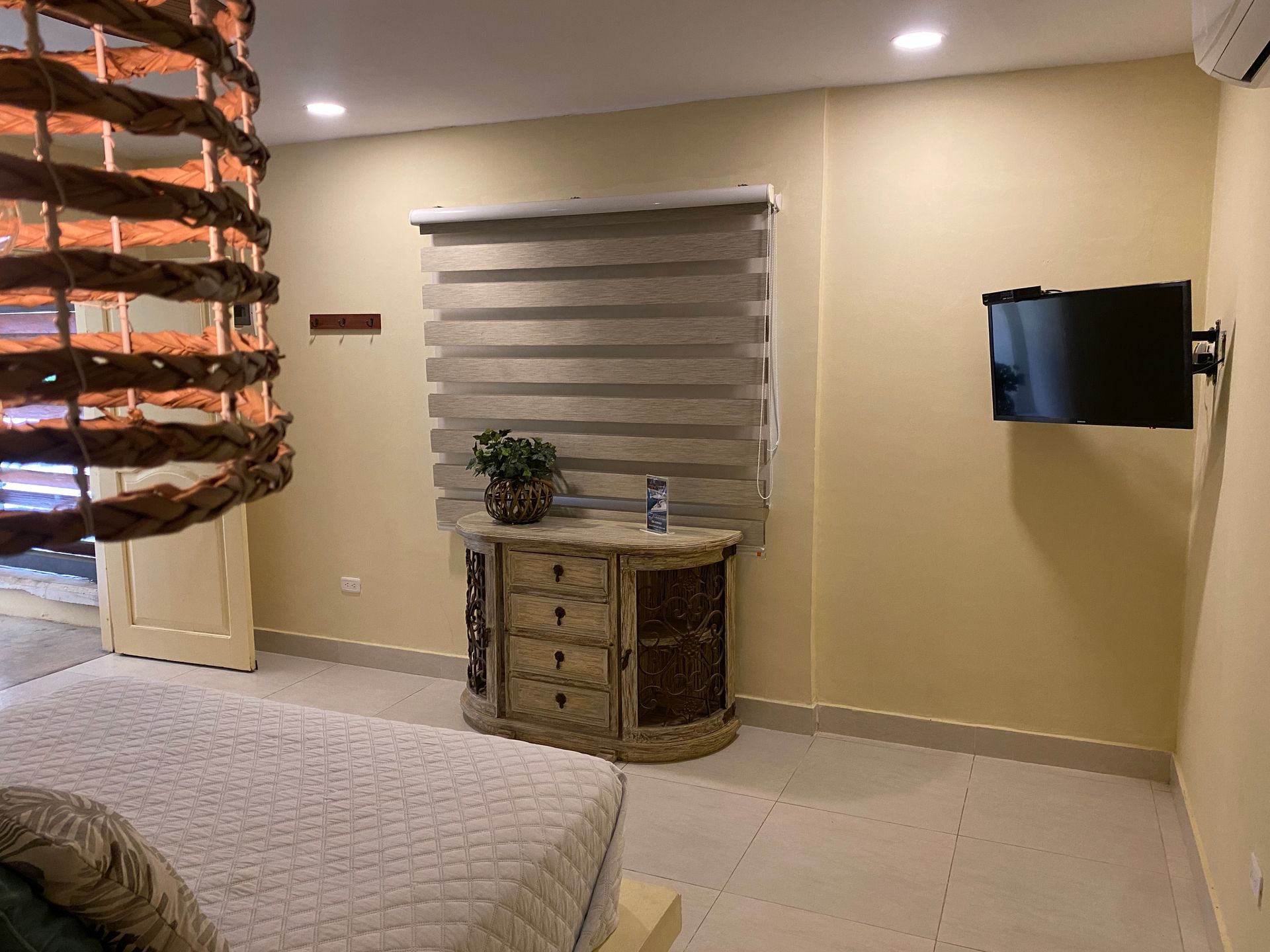 Bedroom interior with bed, dresser, window shades, TV, and decorative hanging.