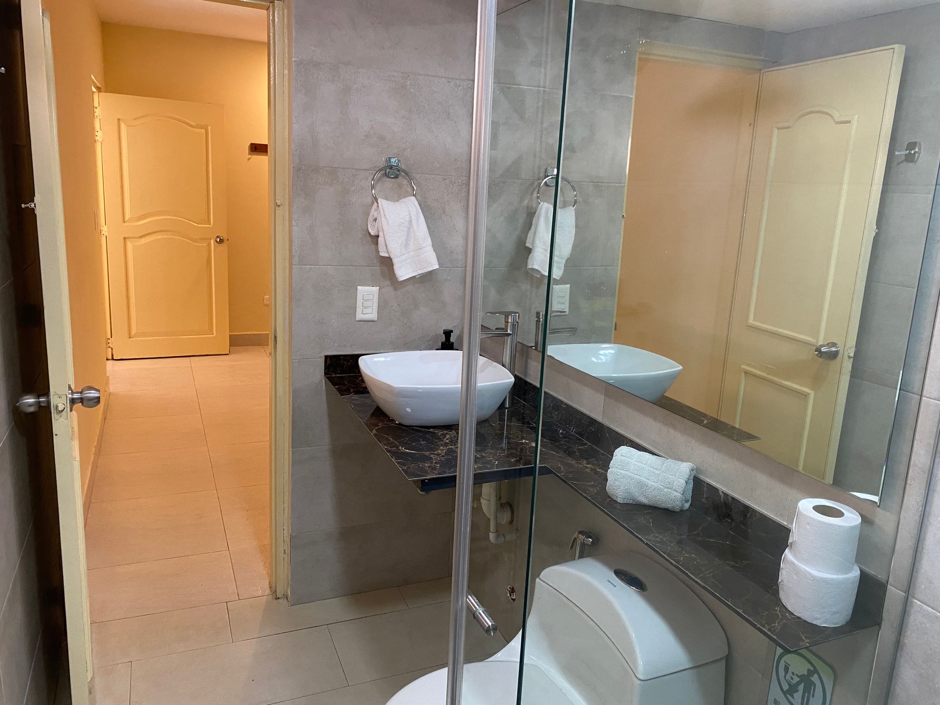 Bathroom with sink, toilet, and mirror, door in the background. White and gray tones.