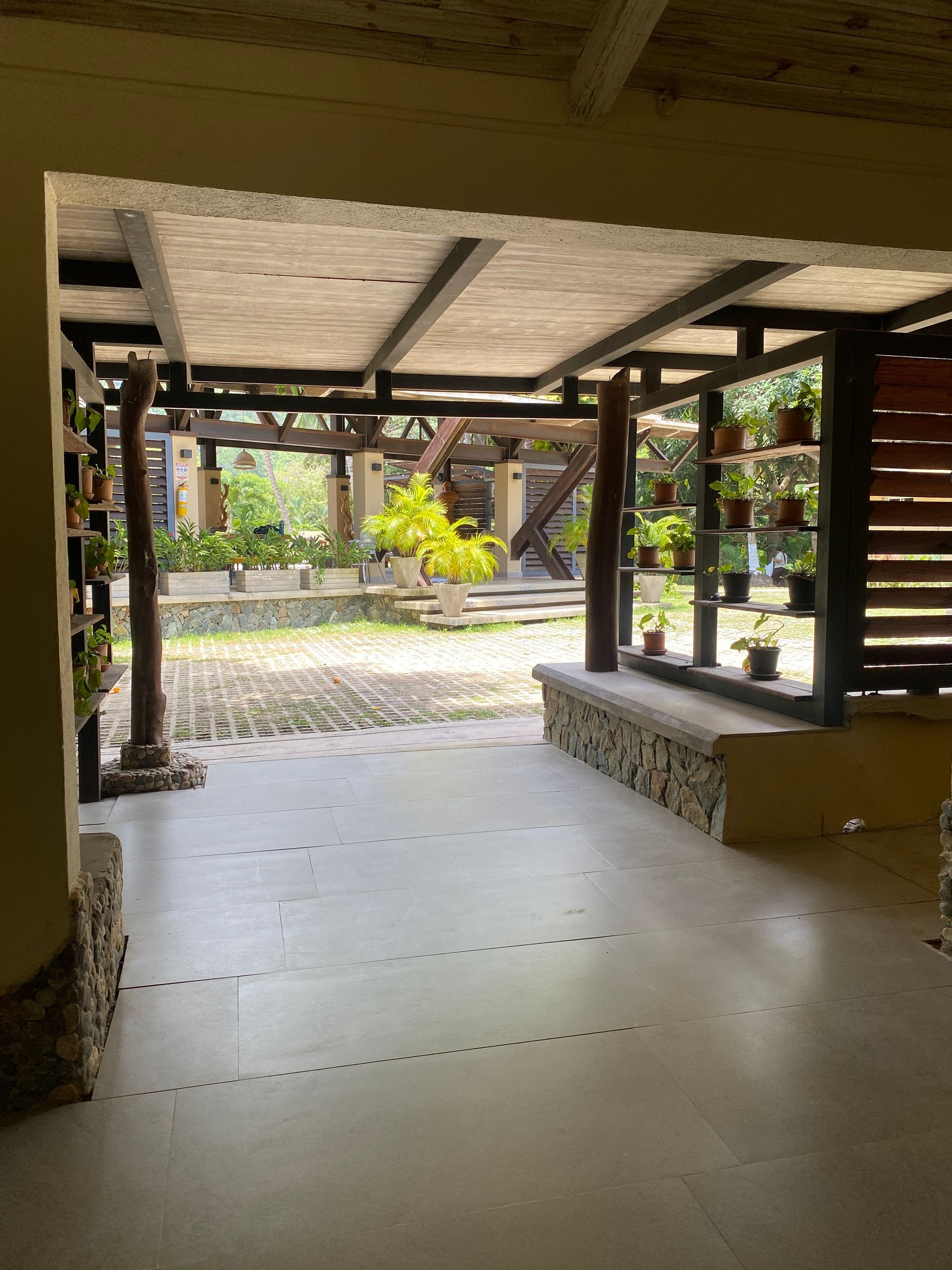Covered patio leads to a garden with plants, stone walls, and wooden beams.