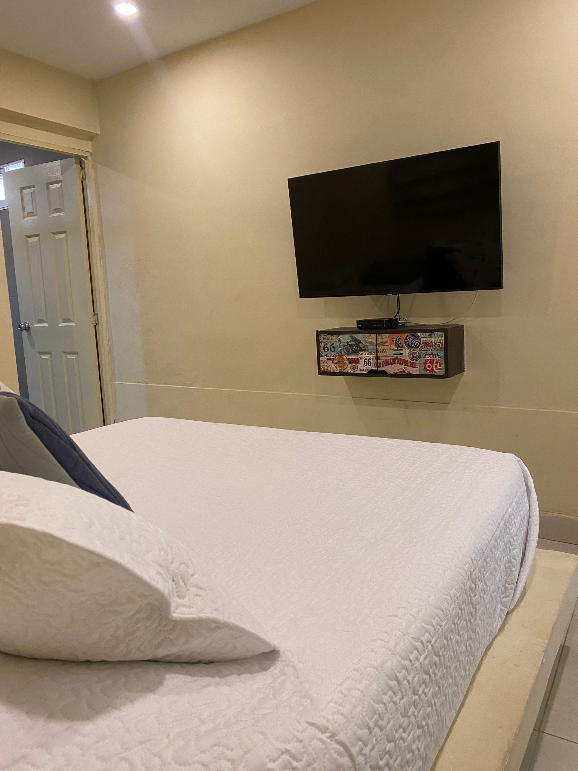 Bedroom with a white bed, TV, and floating shelf mounted on a beige wall.