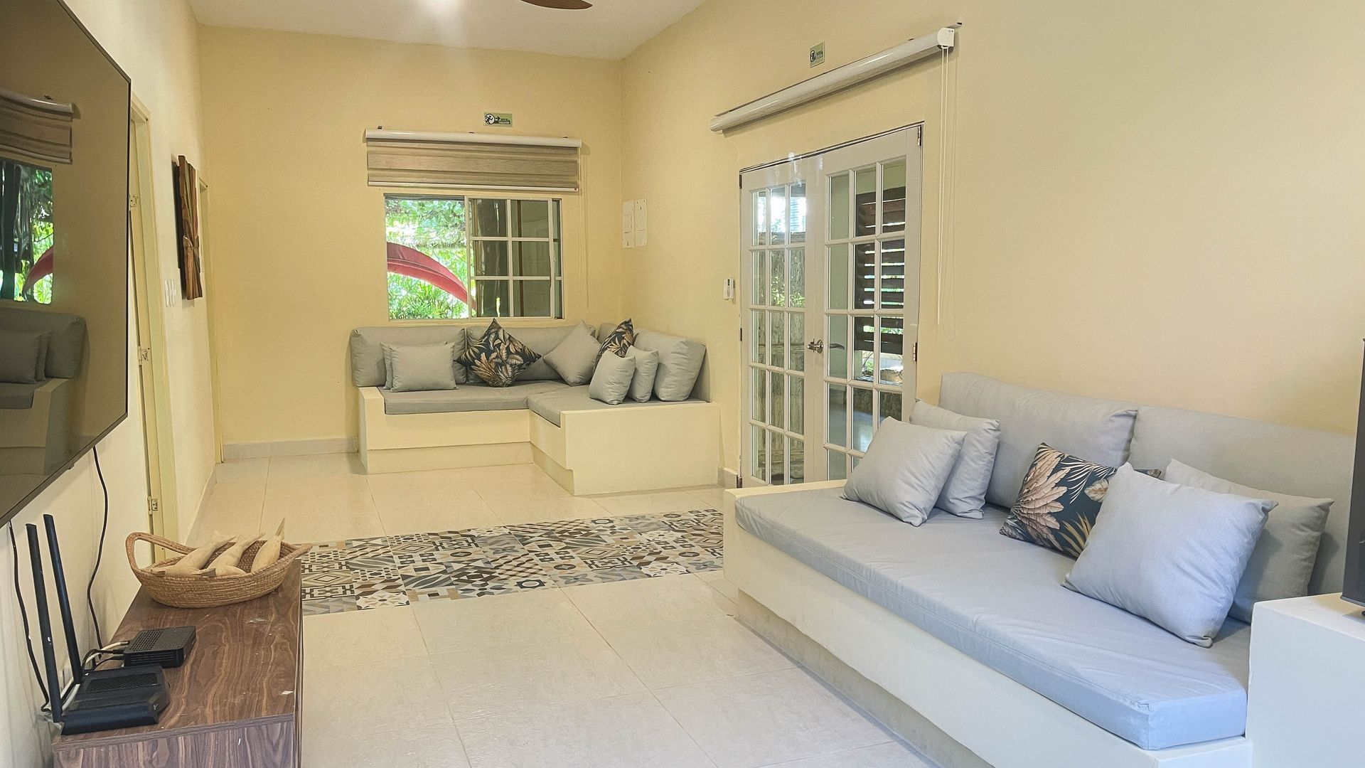 Living room with light-colored walls and built-in sofas with gray cushions. A rug and doors leading outside are visible.