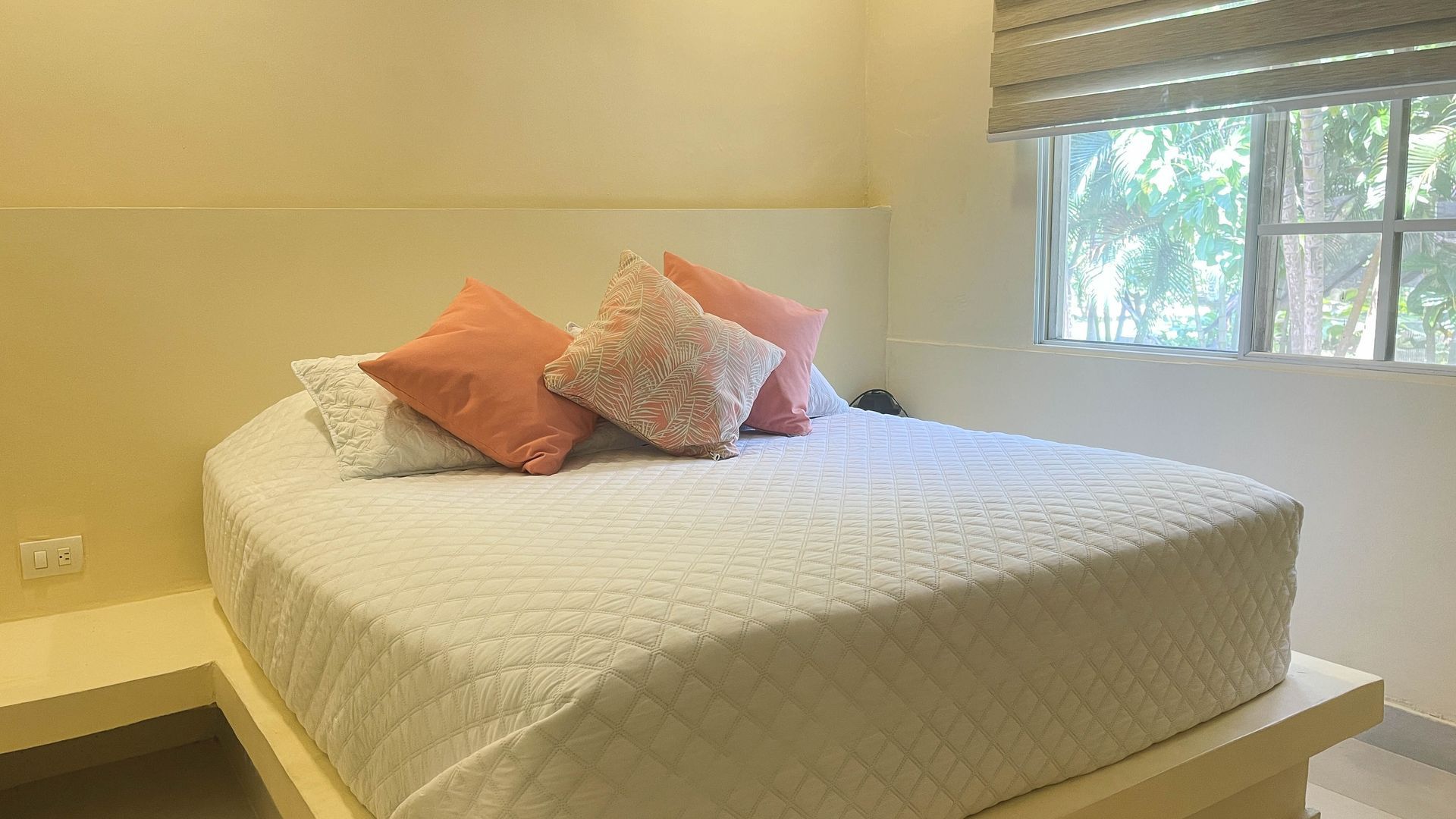 Bed with white quilted cover, coral and white pillows, next to a window with blinds.