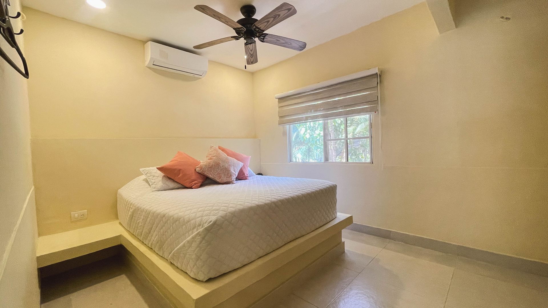 Bedroom with a round bed, window with blinds, ceiling fan, and AC unit. Beige walls and tiled floor.
