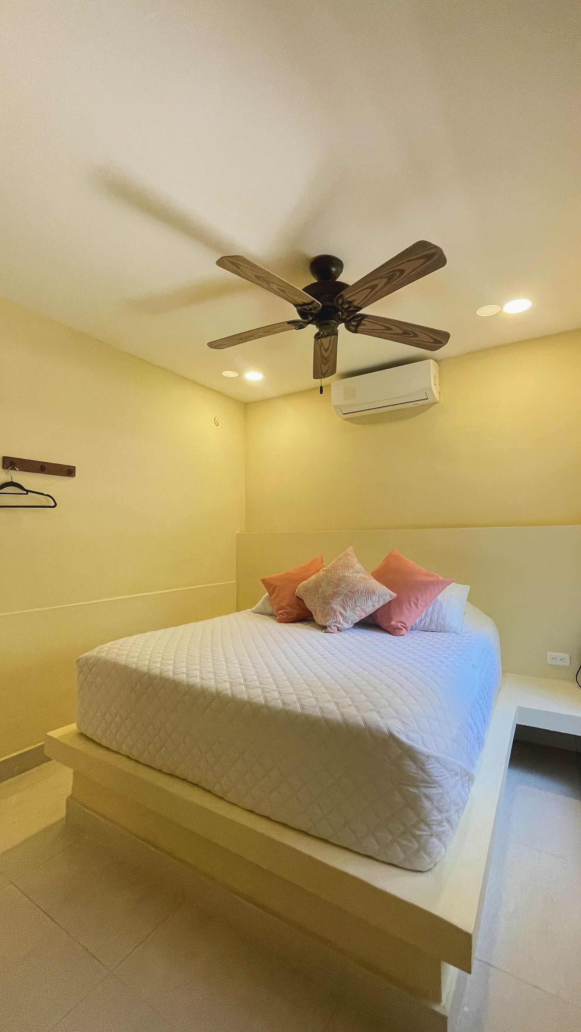 Bedroom with a bed, pillows, ceiling fan, and air conditioner. Walls are beige.