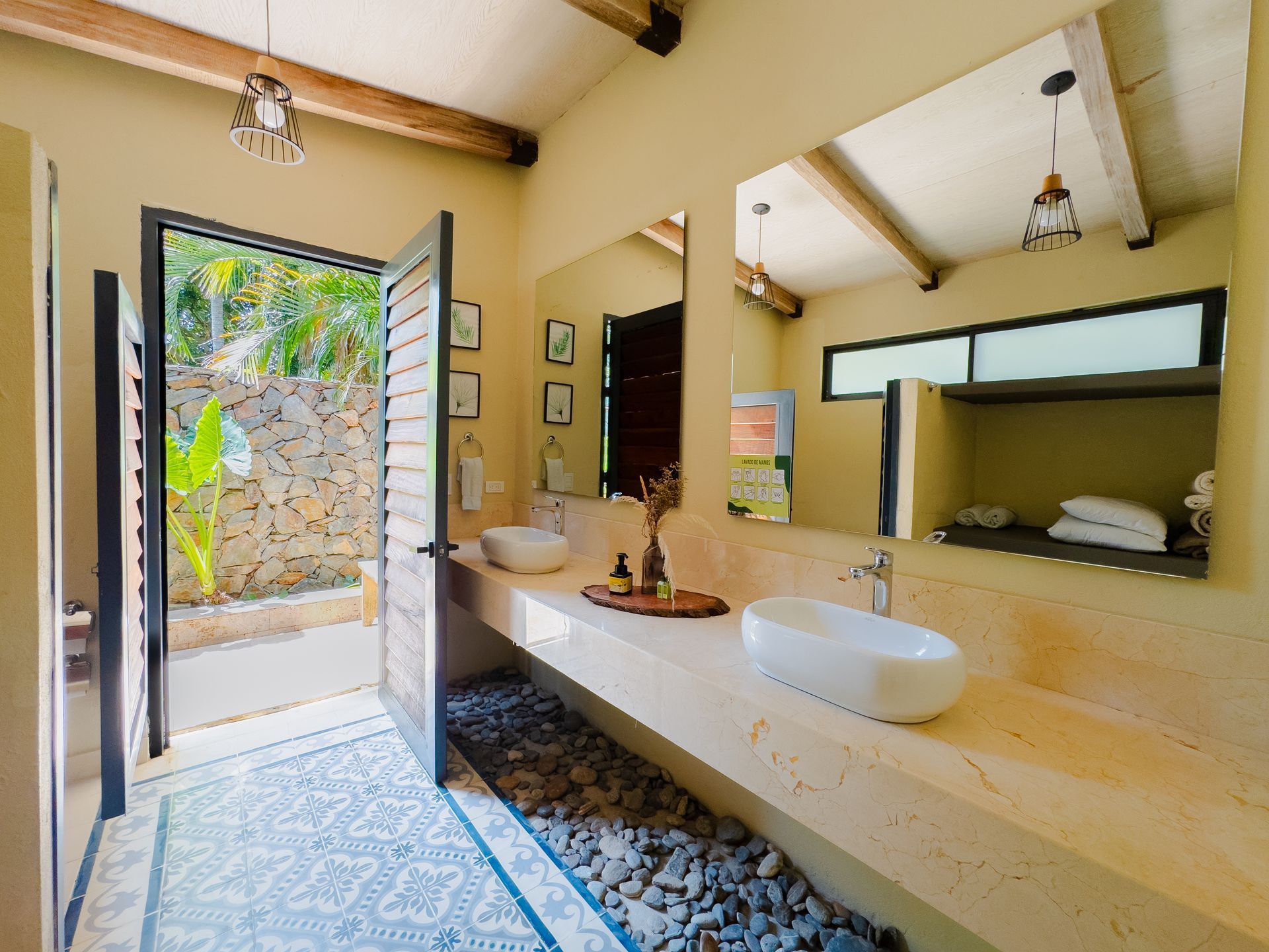 Bathroom with stone wall, wooden beams, open door to garden. Two sinks on a long counter.