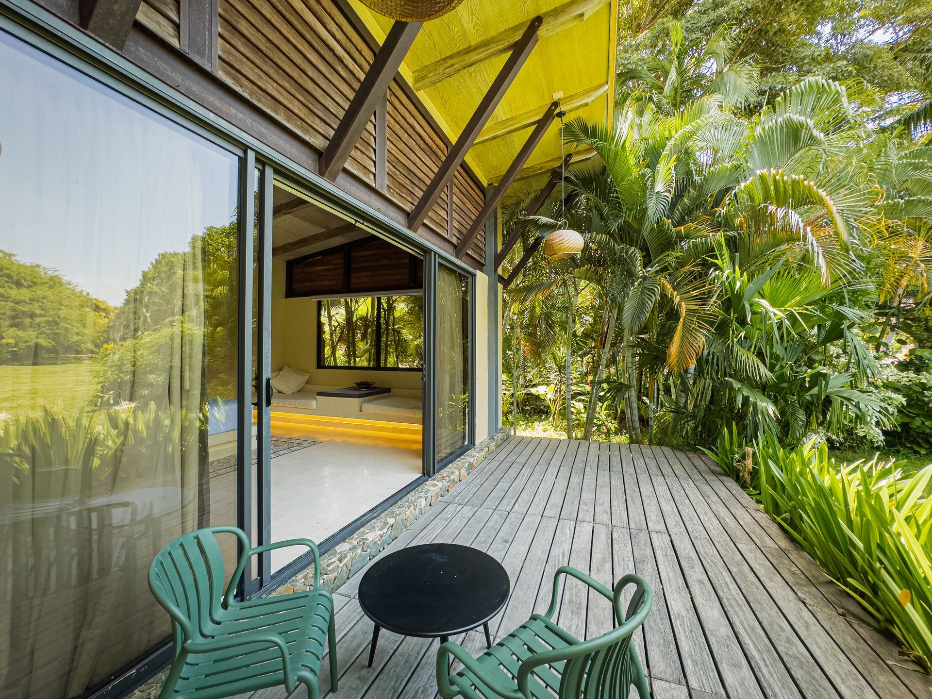 Wooden deck with green chairs, sliding glass door, and view of a bedroom, surrounded by lush greenery.