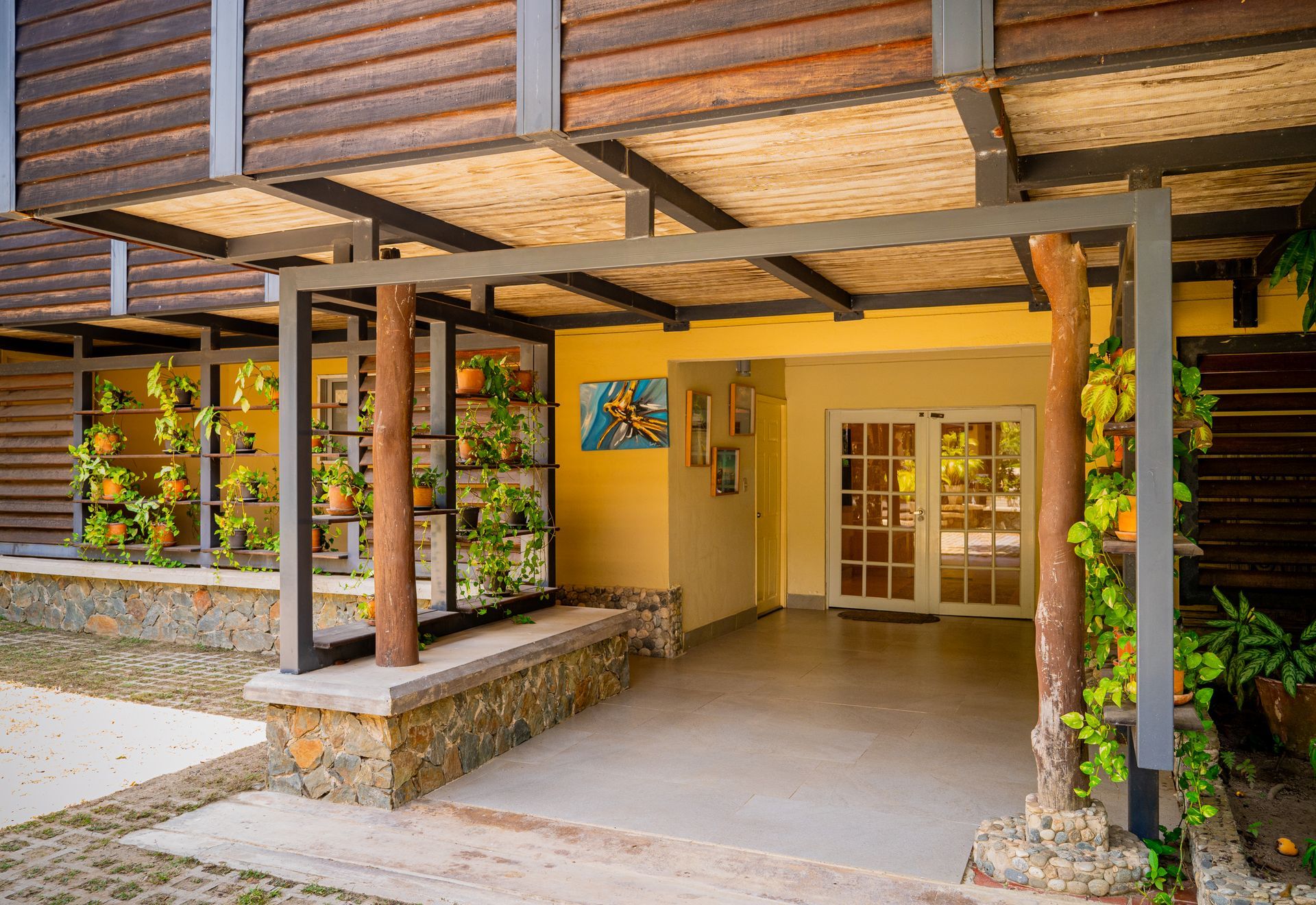 Covered porch with trellis, plants, and entrance to a building. Yellow walls, stone and wood details.