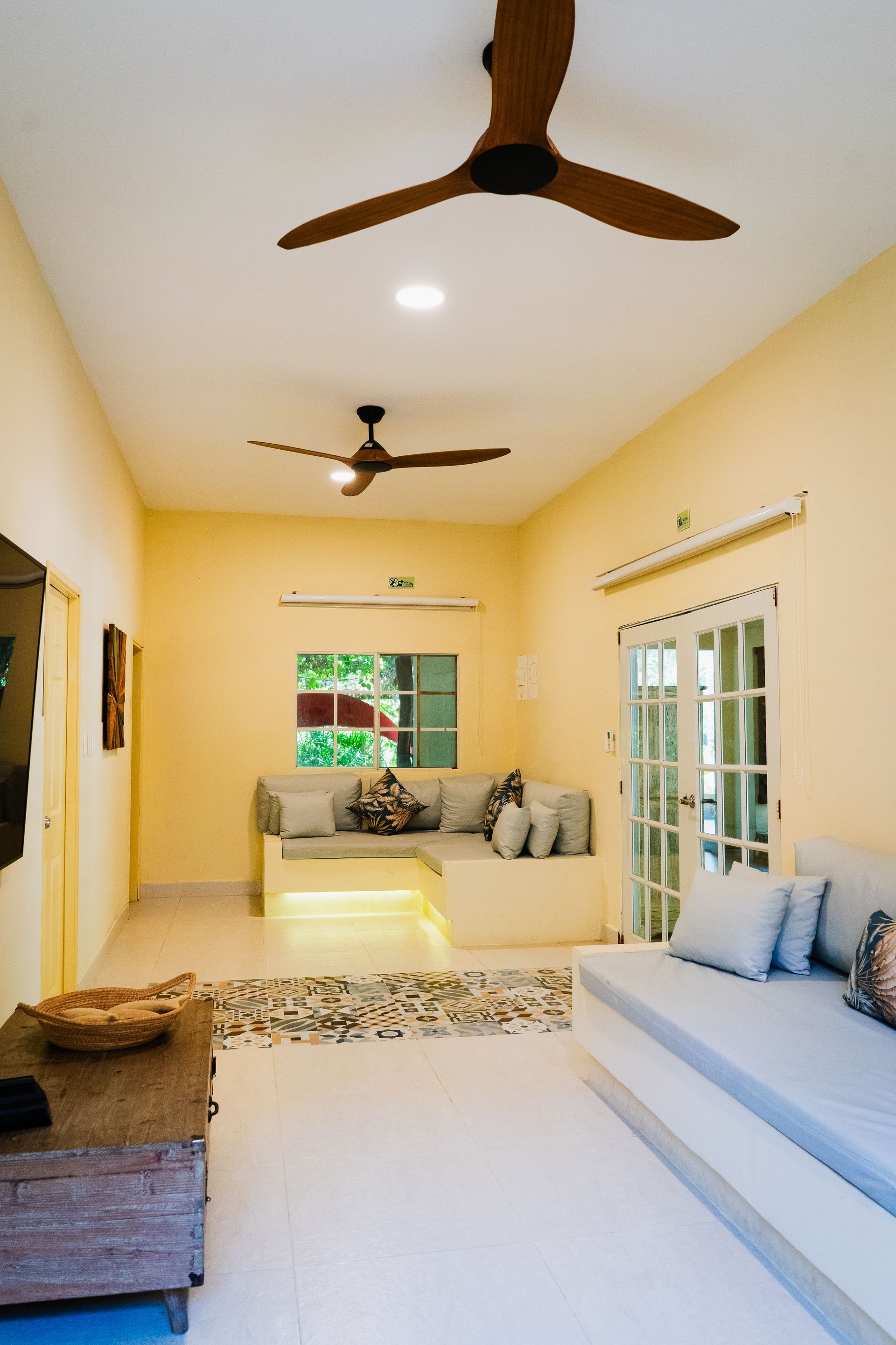 Bright living room with two ceiling fans, built-in seating, and a window. Light yellow walls and white flooring.