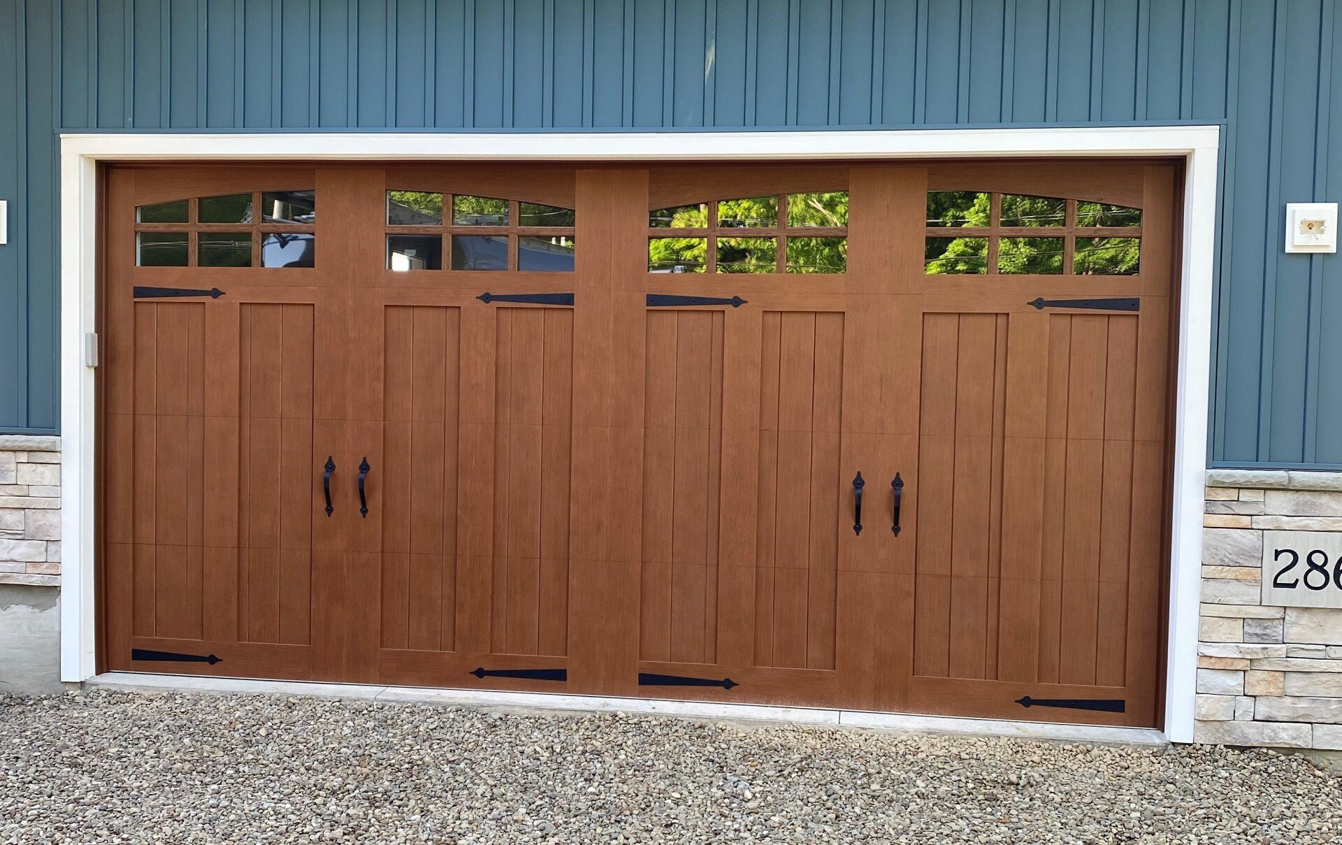 Brown Craftsman-style garage door with windows and black hardware; set against blue siding and gravel.