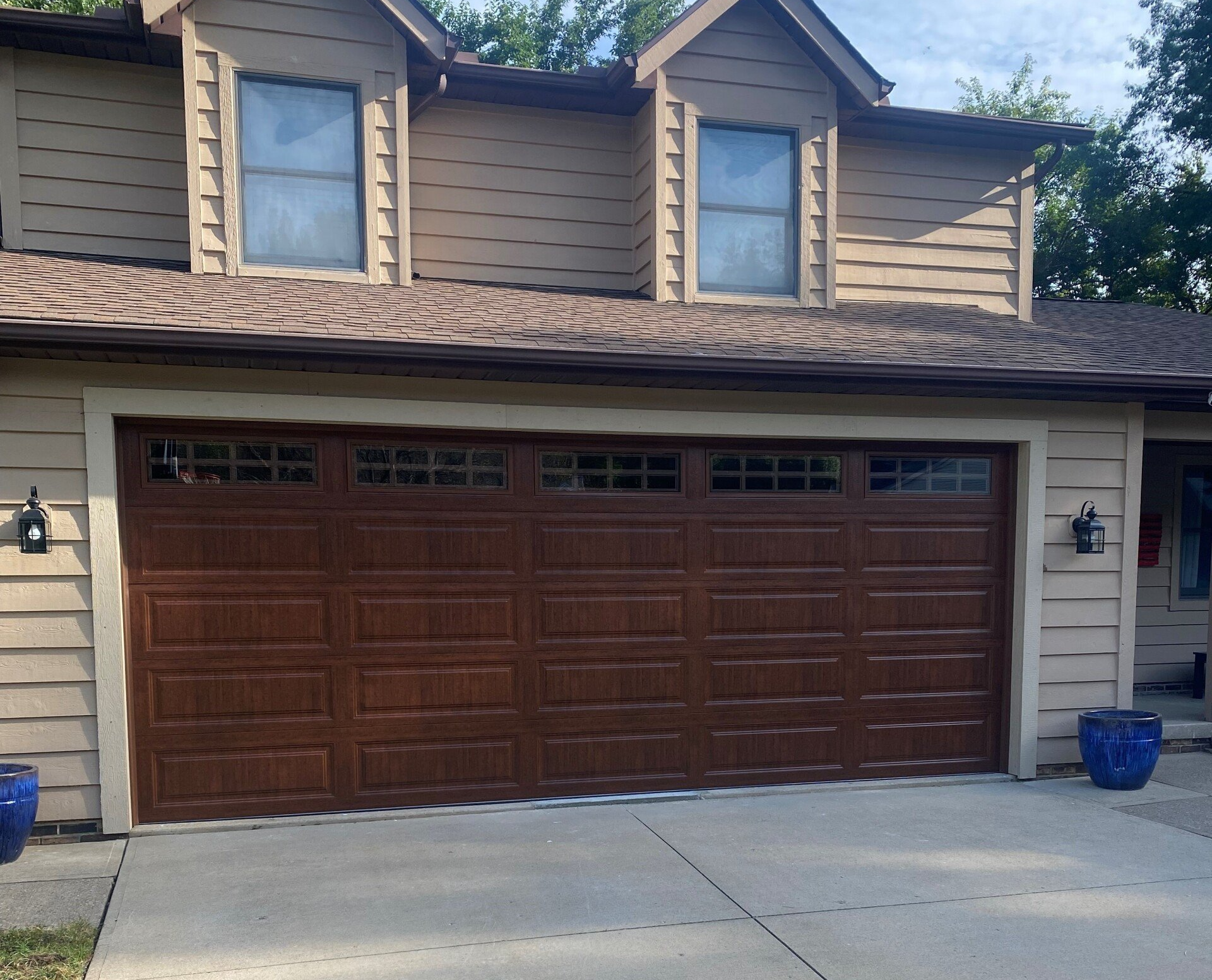 Brown garage door on a two-story house with two dormers. Tan siding, dark brown roof.
