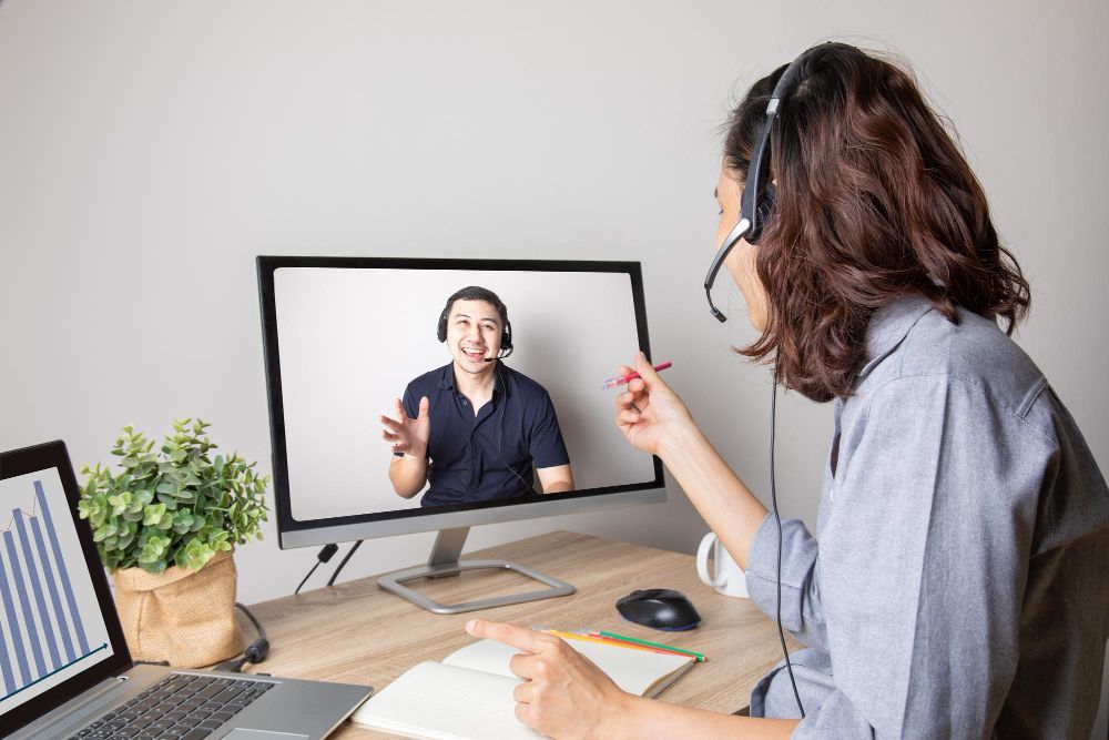 A woman wearing headphones is having a video call with a man on a computer screen.