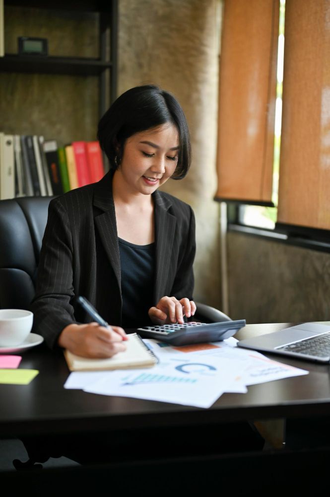 A woman is sitting at a desk using a calculator and writing in a notebook.