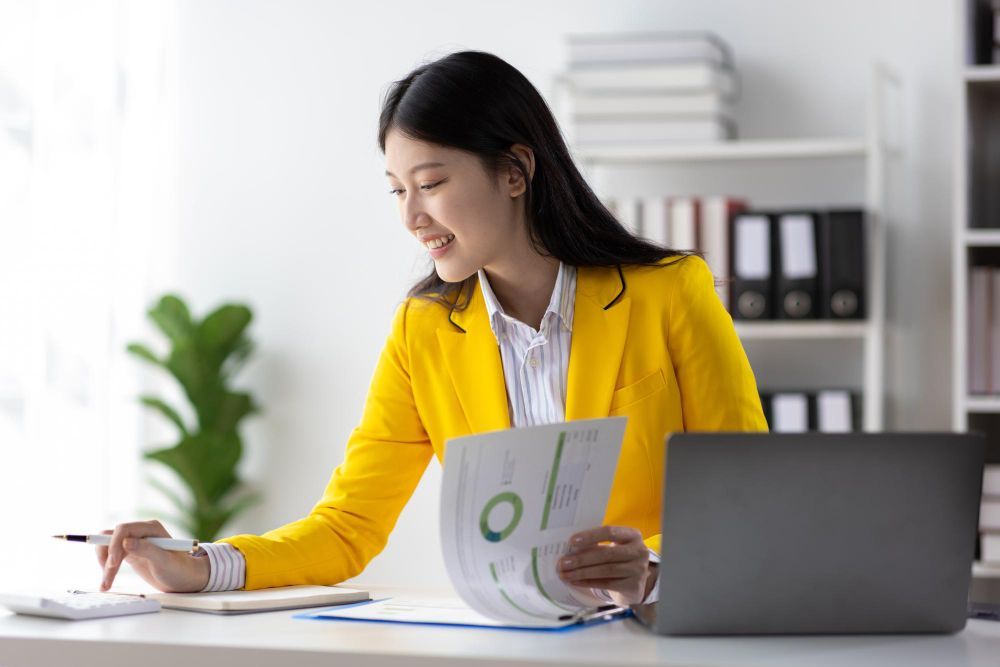 A woman in a yellow jacket is sitting at a desk with a laptop and a calculator.
