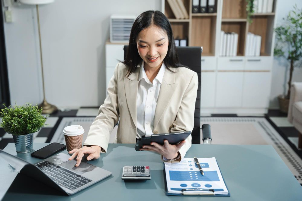 A woman is sitting at a desk using a tablet and a laptop.