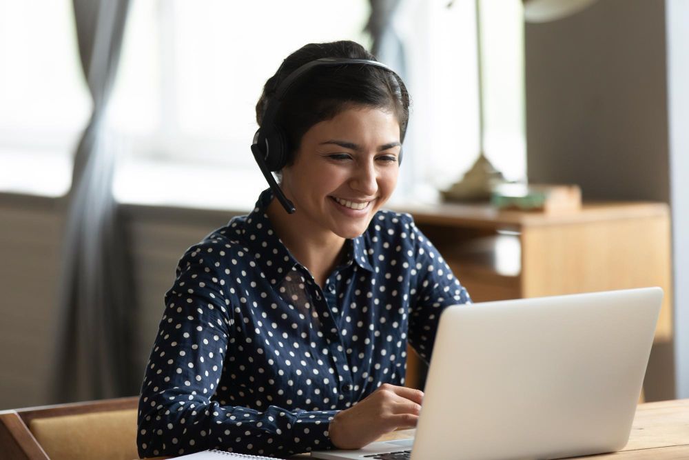 A woman wearing headphones is sitting at a table using a laptop computer.