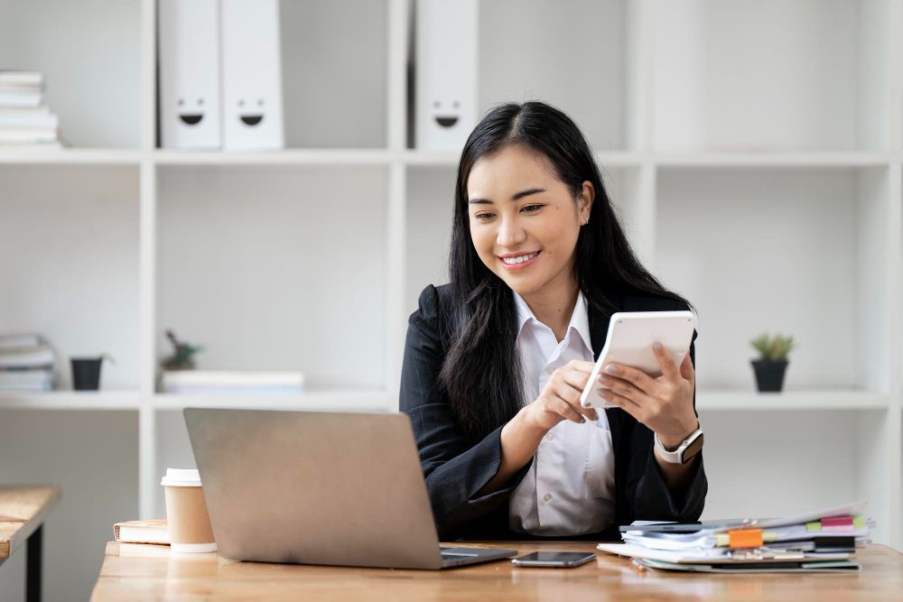 A woman is sitting at a desk using a laptop and a tablet.
