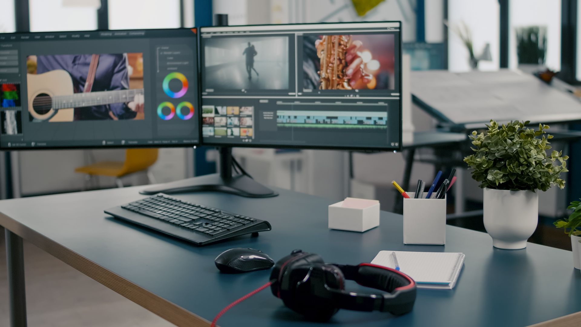 Desk with dual monitors displaying video editing software, headphones, keyboard, and a plant.
