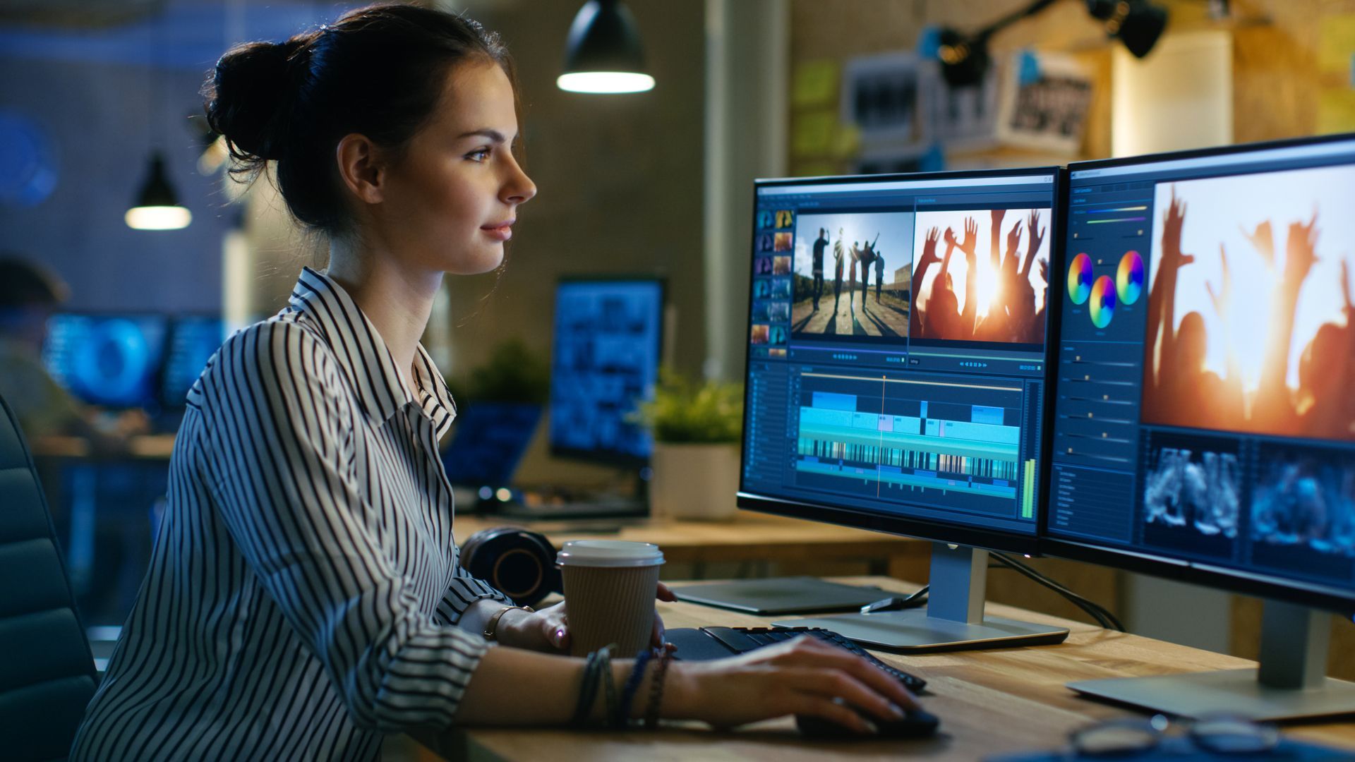 Woman at computer editing video, with dual monitors displaying footage. Office setting.