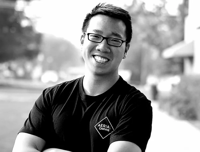 A man wearing glasses and a black shirt is smiling in a black and white photo.