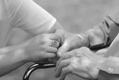A black and white close-up shows a person holding the hands of an older person sitting in a wheelchair.
