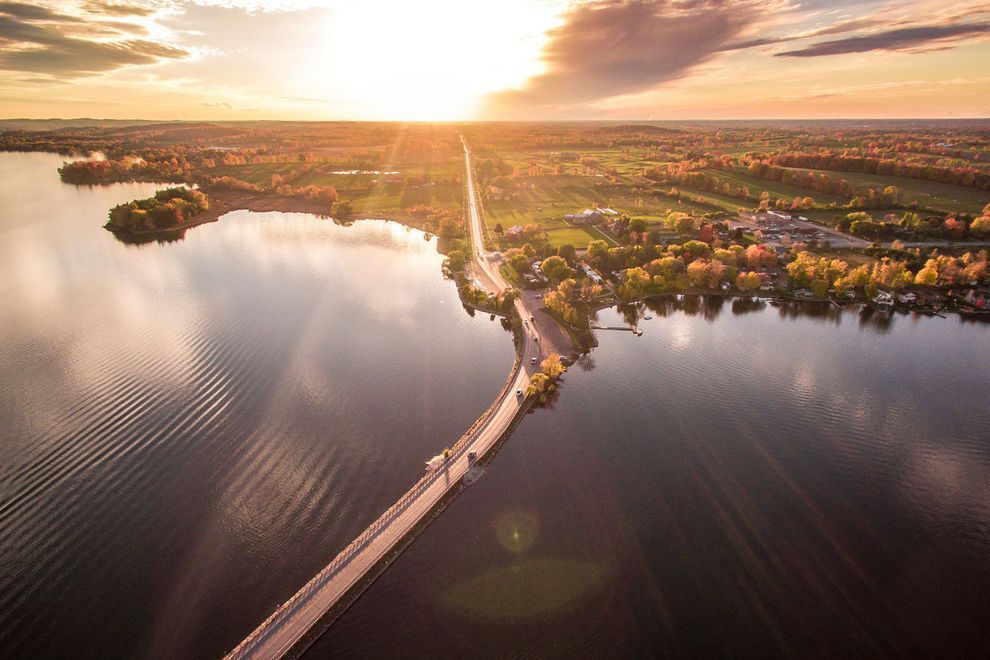 Bridge over calm water at sunset, road leads to horizon, orange and yellow sky, trees and buildings along the shores.