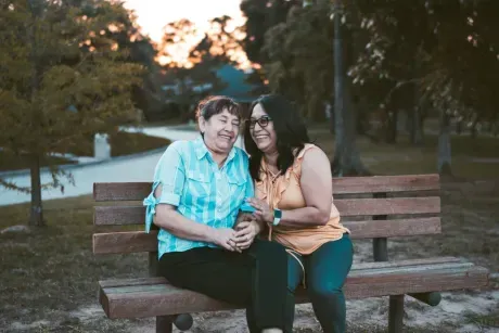 Two smiling people sit on a wooden bench in a park during dusk, engaging in conversation.