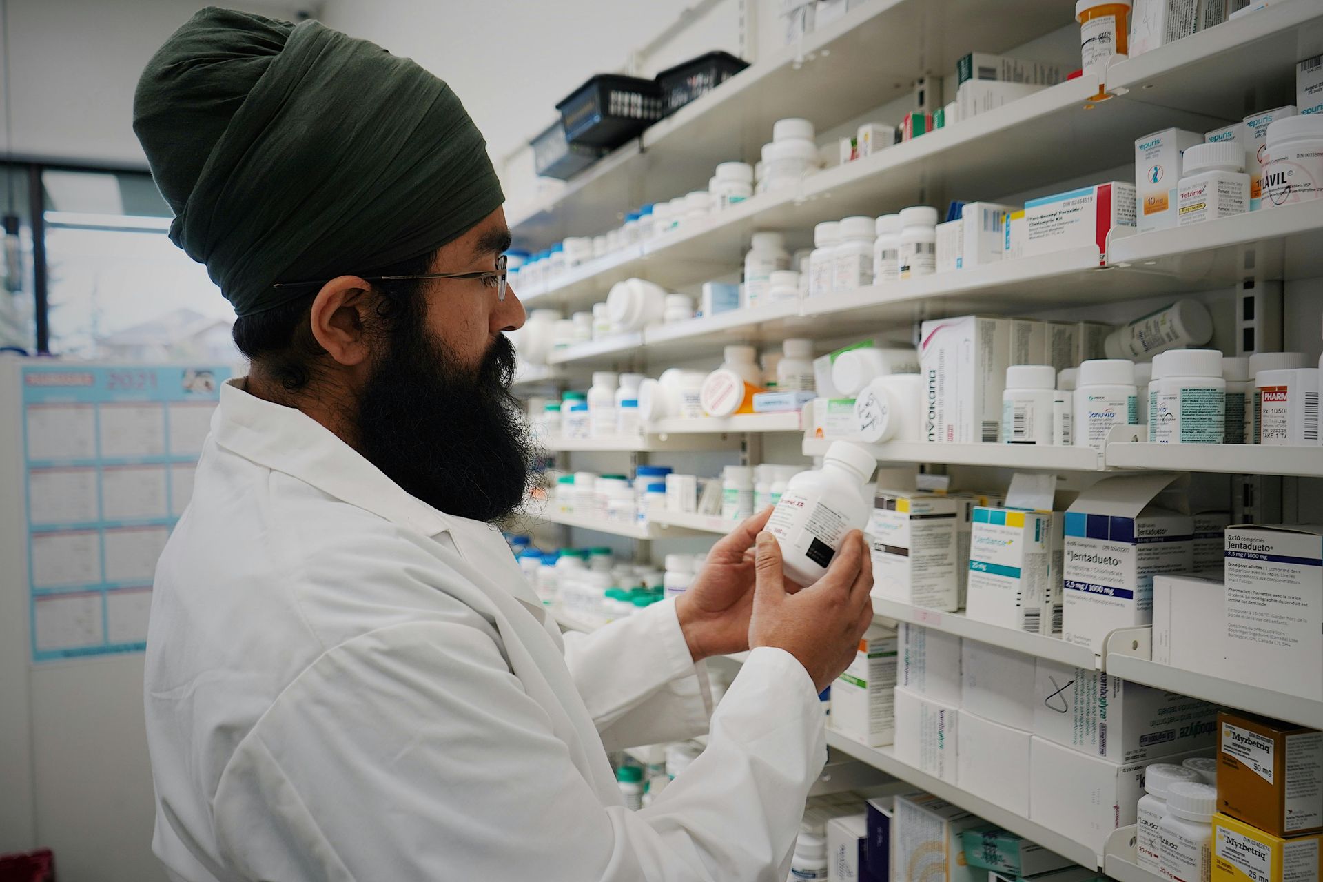 A pharmacist wearing a lab coat and green turban holds a medication bottle, checking a shelf in a pharmacy.