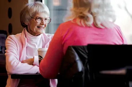 Two people sitting at a table in a cafe, facing each other and smiling while having a conversation over coffee.
