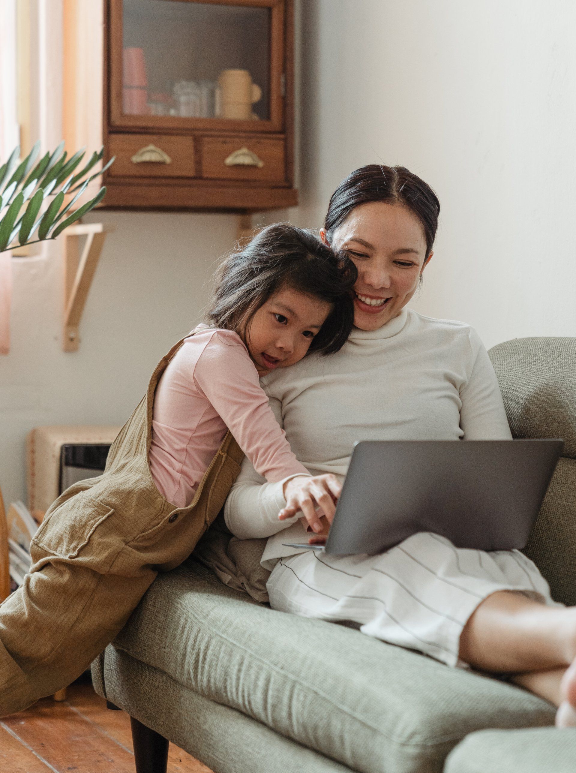 A smiling adult and child on a couch looking at a laptop screen together in a bright, homey living room.