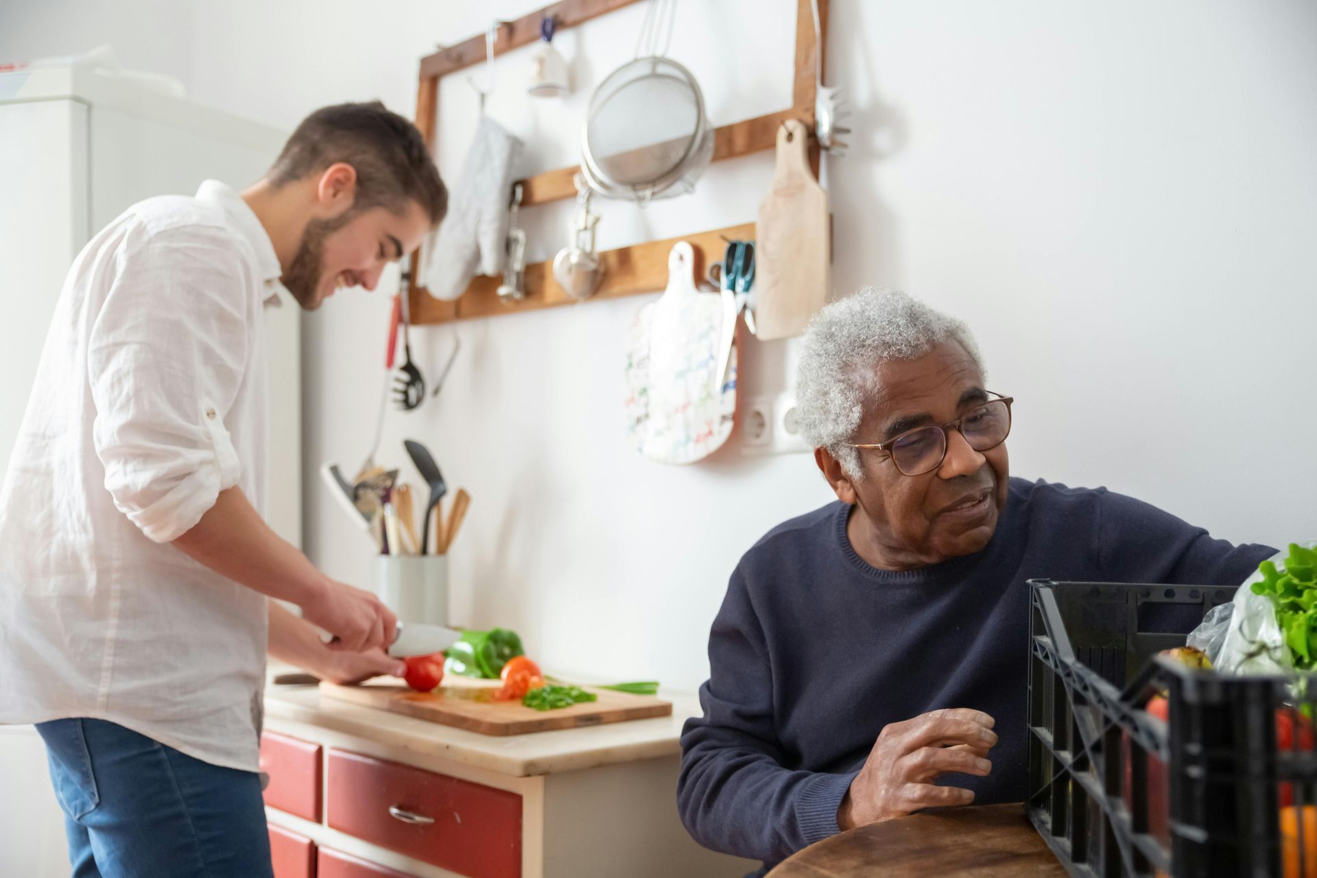 Young person cutting vegetables; older person reaches into a black crate in a kitchen.