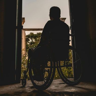 A silhouette of a person sitting in a wheelchair, facing a doorway that opens to a view of trees and sky.
