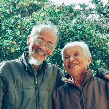 Two smiling individuals in dark zip-up jackets standing together outdoors in front of a leafy green bush.