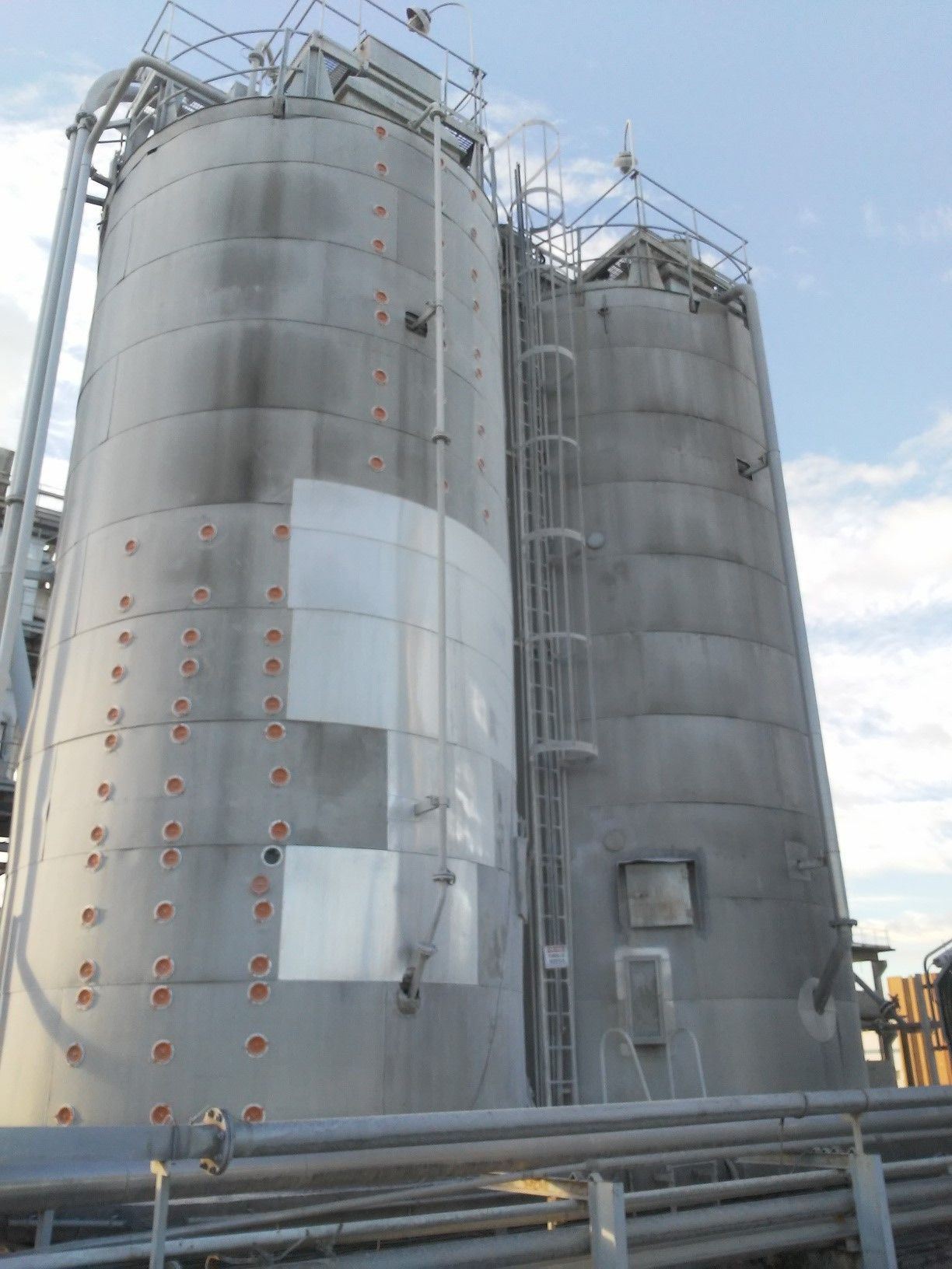 Two tall gray industrial silos with ladders and metal infrastructure against a blue sky.
