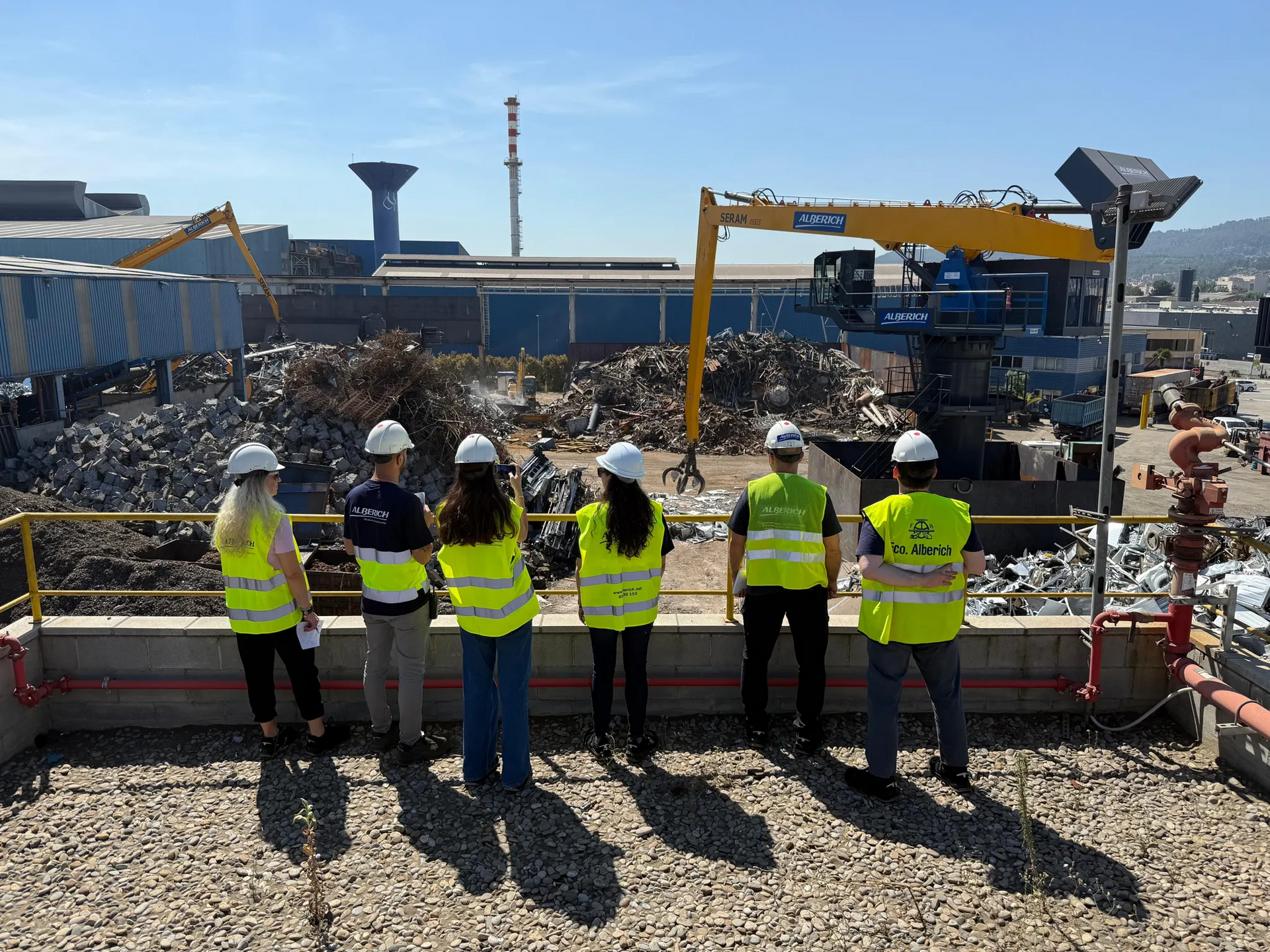 Group of people in safety vests and helmets, viewing a scrap metal yard on a sunny day.
