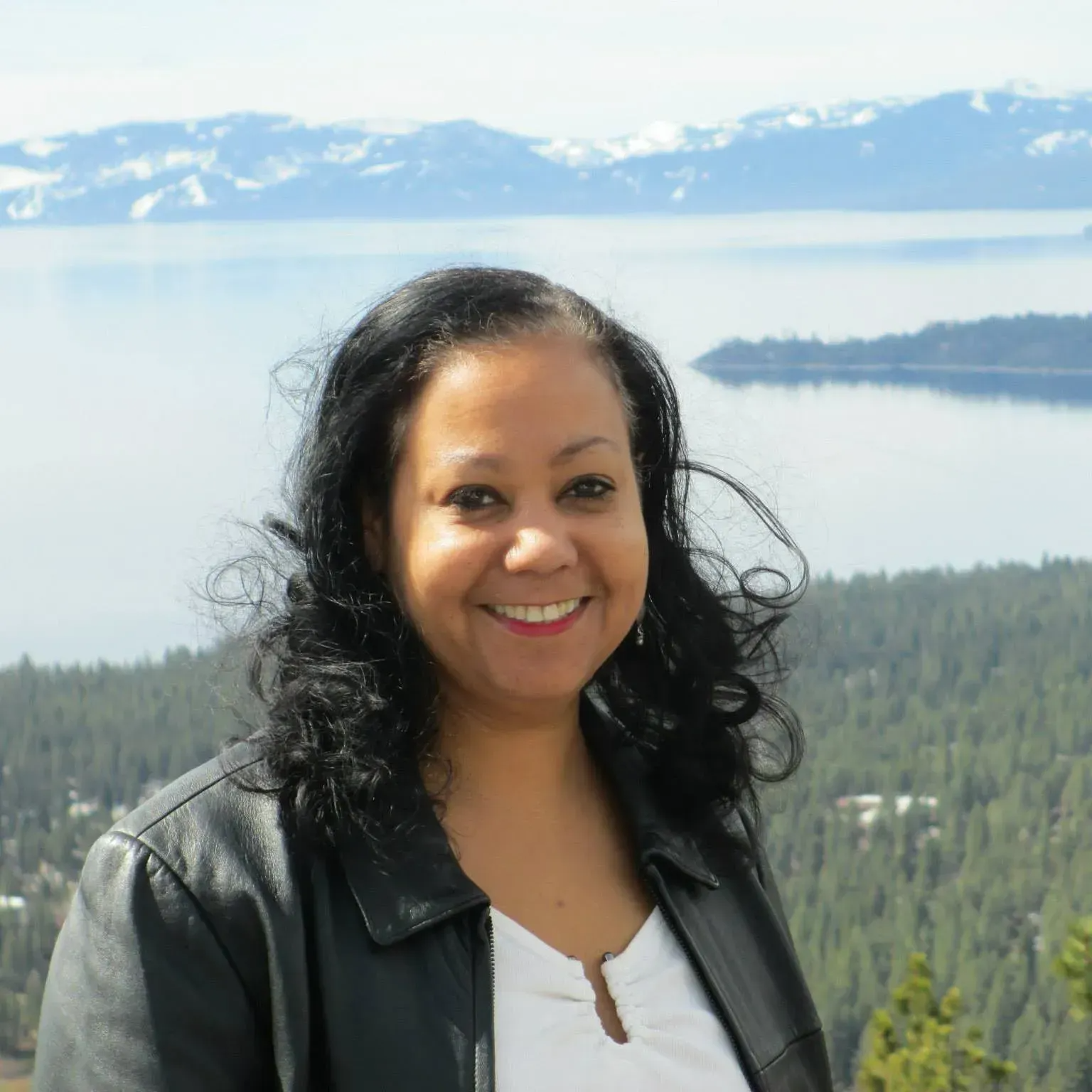 A woman in a black leather jacket smiles in front of a lake