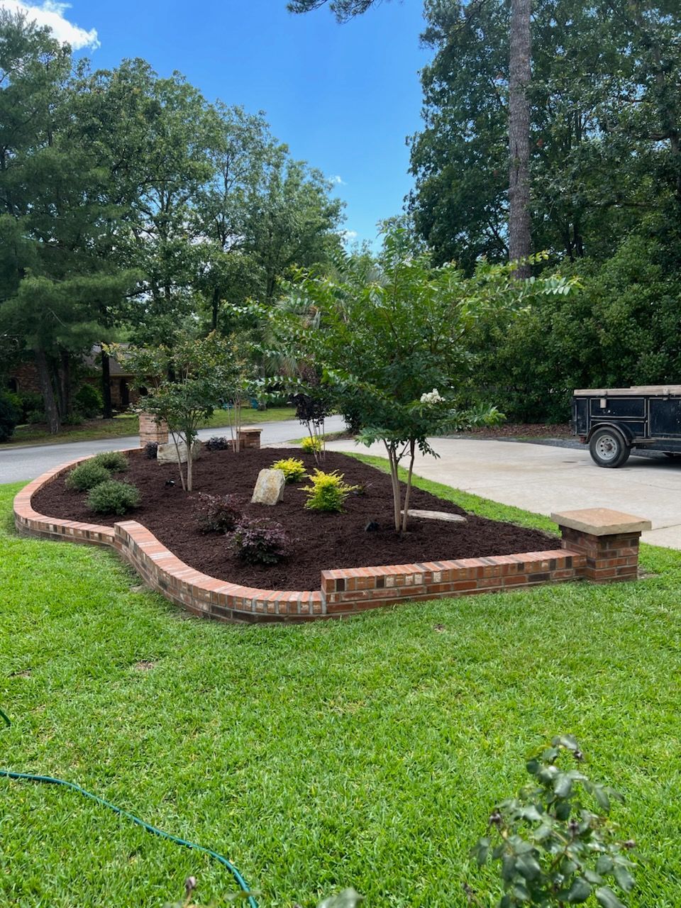 Brick-edged flower bed with mulch, shrubs, and small trees on a grassy lawn with a driveway and trees in the background.