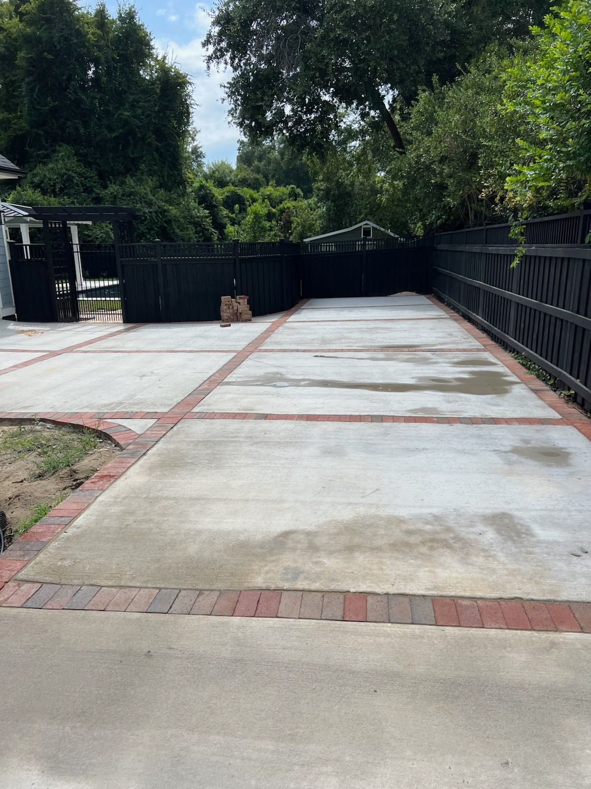 Concrete driveway with red brick border, black fence, and trees.