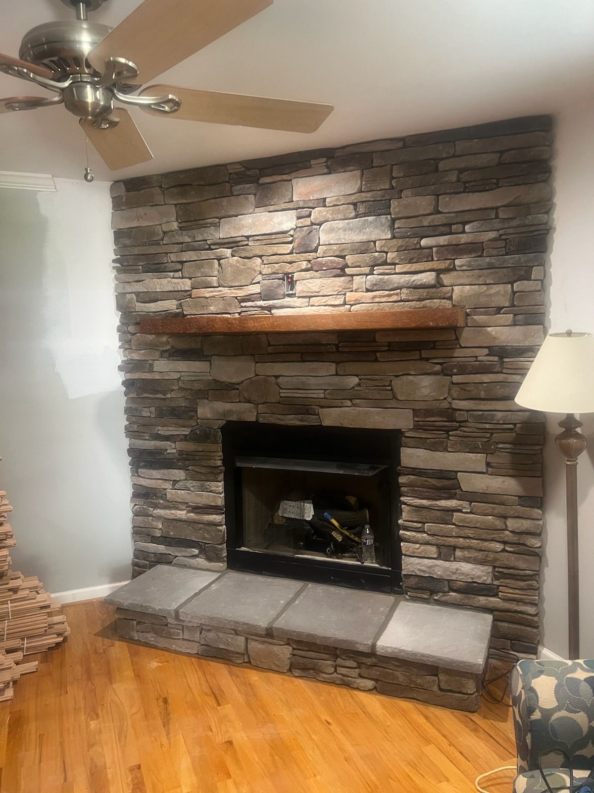 Stone fireplace with a wooden mantle and hearth, flanked by a lamp and ceiling fan.