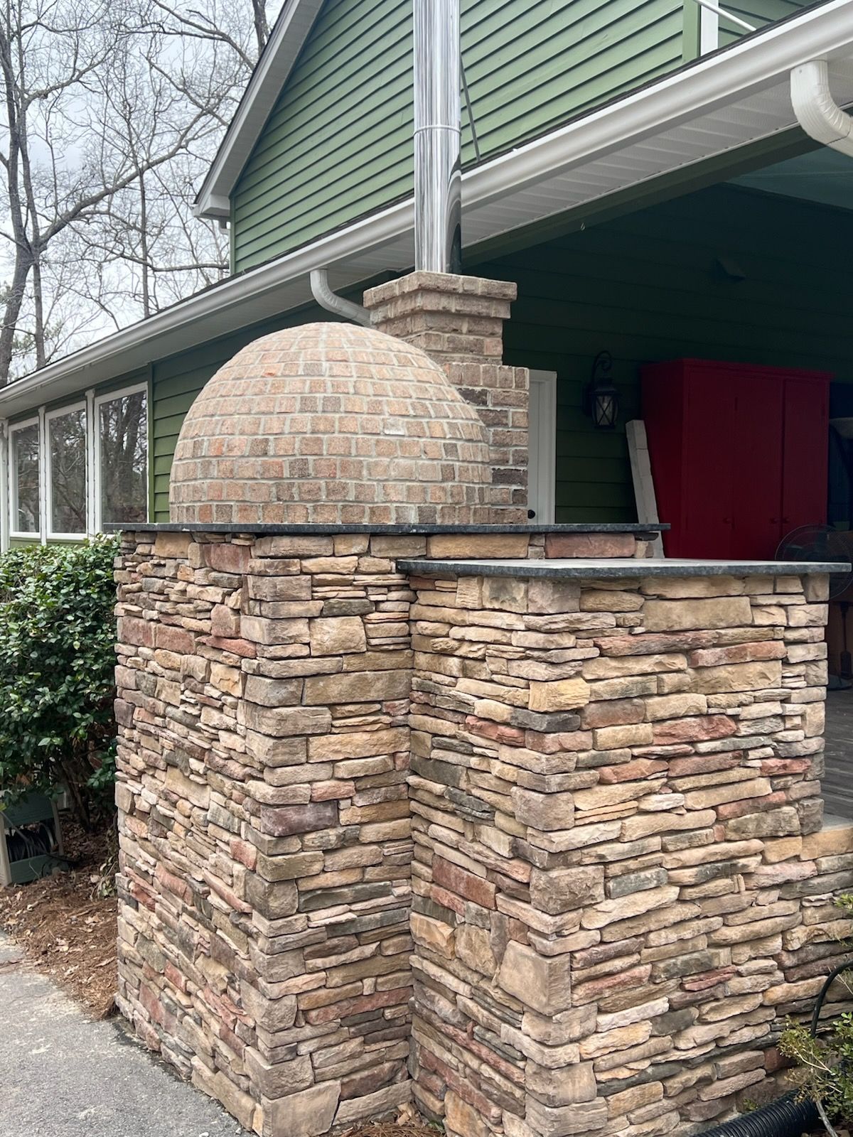Outdoor brick pizza oven with stone base, chimney, and green siding in the background.