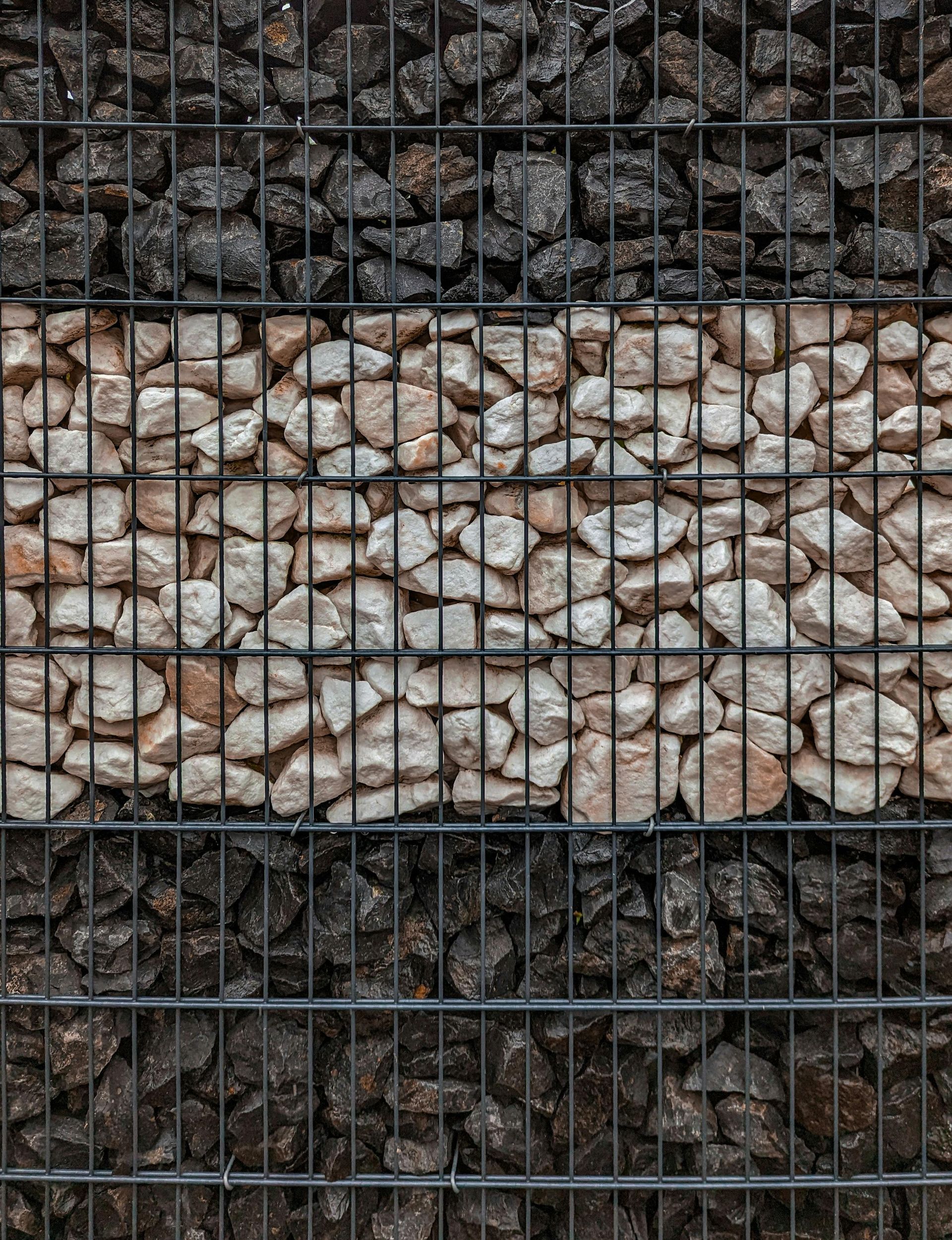 A gabion wall section with black rock top and bottom, separated by tan rock in the center.
